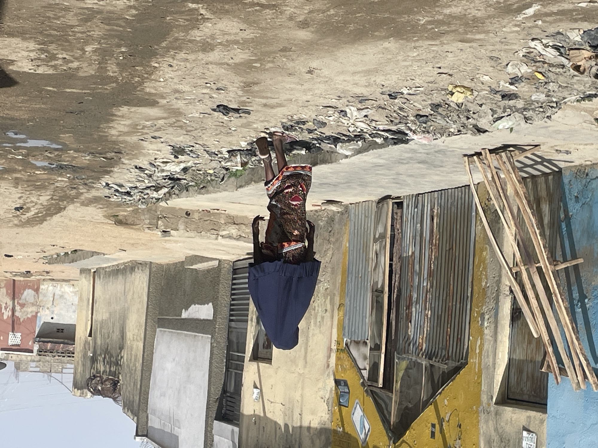 A street scene in what appears to be a densely populated urban area, potentially a slum or impoverished neighborhood. The street is littered with debris, trash, and what looks like dried mud or cracked pavement. Buildings on either side have a weathered, run-down appearance, with corrugated metal and mismatched paint. A person in dark clothing and a blue skirt is visible, possibly a woman, but their face is not clearly seen. The overall impression is one of poverty and neglect.