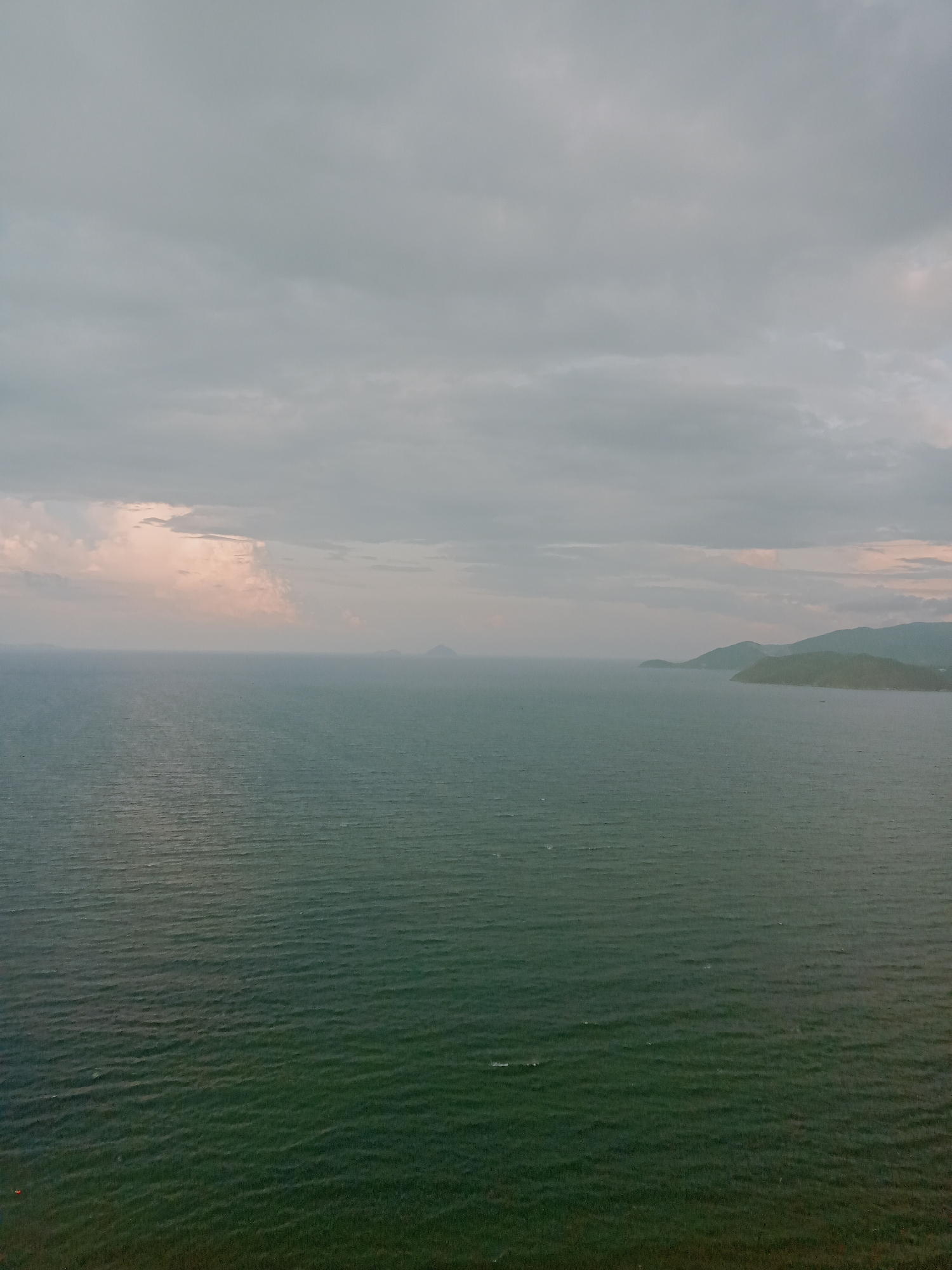 The image presents a wide view of the sea, taken from an elevated perspective. The sky is overcast with gray clouds, suggesting a cloudy or potentially rainy day. A faint light color appears near the horizon.
The ocean surface is a dark green, with subtle waves creating texture across the water. In the distance, landmasses are visible; a smaller island or mountain is present to the center-left and a larger coastline is on the right.
Based on location of Nha Trang Ward, Vietnam, this image is possibly looking from the shore out over the ocean towards the several islands on the coast.