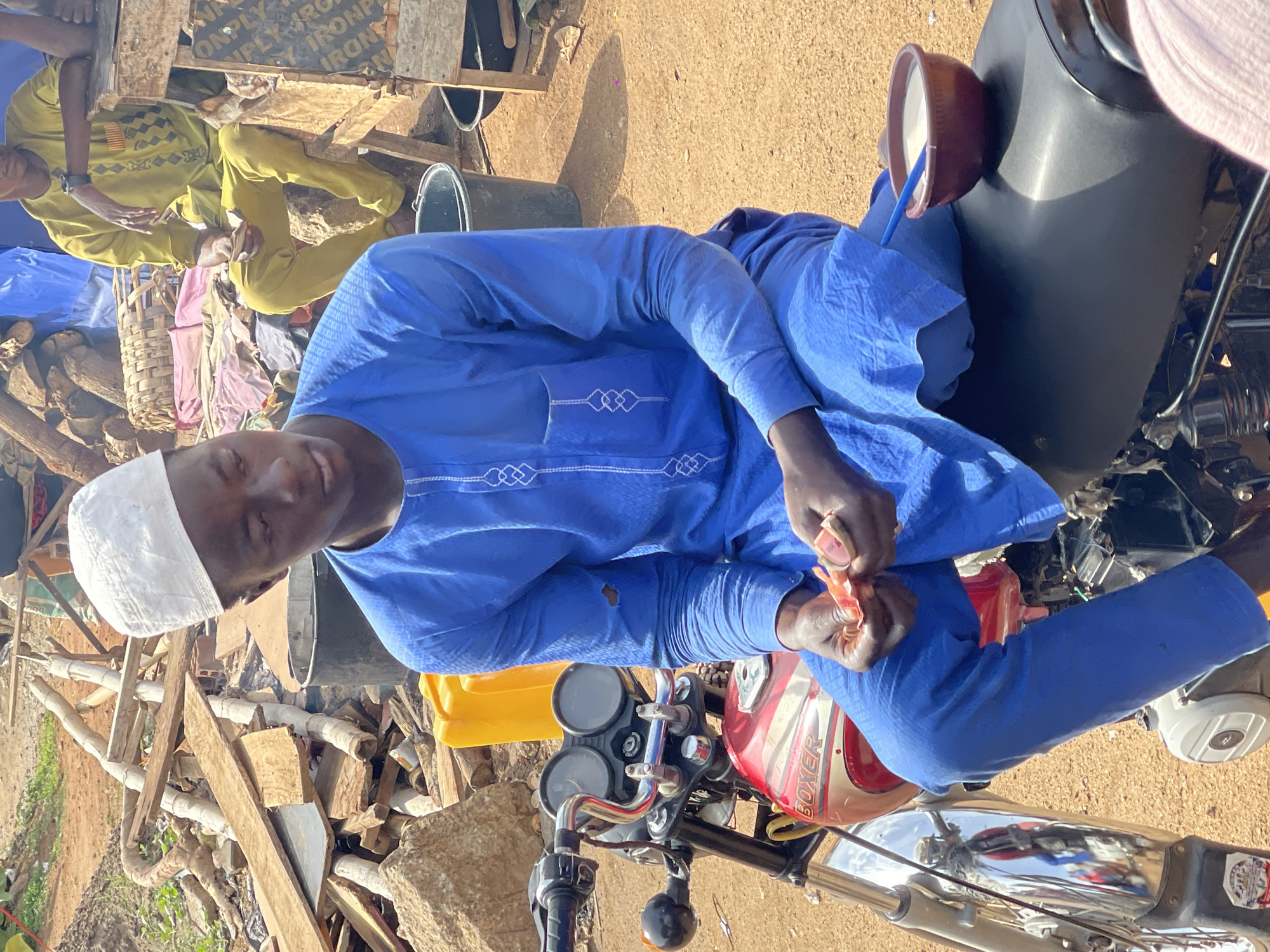 A man in a blue tunic and white kufi cap is seated on a red and white Boxer motorcycle, holding small red objects, presumably food, in his hands. A bowl with white liquid is perched on the motorcycle behind him. The man is situated amidst a somewhat cluttered outdoor environment with wood and various objects scattered around. Another person, dressed in yellow, sits in the background. The location is likely a roadside or a market area in Gwarinpa, Nigeria, given the setting and the man's attire.
