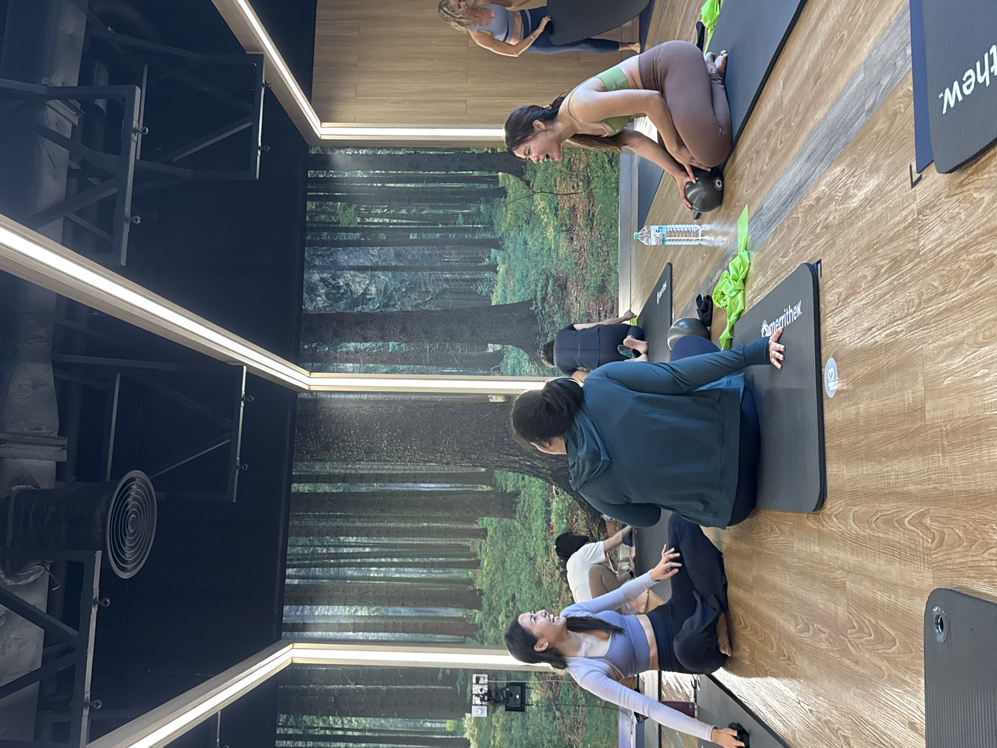In a dimly lit yoga studio with a forest mural as a backdrop, several individuals are seen participating in a fitness class. The floor is made of polished wood, and the lighting fixtures are modern and integrated into the ceiling.
In the foreground, two women are seated on yoga mats, facing away from the camera. One is wearing a teal hooded sweatshirt and dark pants, while the other is dressed in a green sports bra and brown leggings. Both appear to be engaged in conversation or listening to an instructor.
To the left, a woman in a light purple sports bra and dark leggings is also seated on a mat, facing towards the other women. She is smiling and appears to be interacting with them. Behind her, another person is crouched down on a yoga mat.
In the background, to the right, a woman with blonde hair, wearing a blue sports bra and dark leggings, is standing and holding a black yoga mat. Other participants are visible in the mid-ground and background, also engaged in various poses or preparations.
A water bottle and green resistance bands are scattered on the mats. The overall atmosphere suggests a group fitness session in progress. The location is specified as the city of Nong Pa Khrang, Thailand.