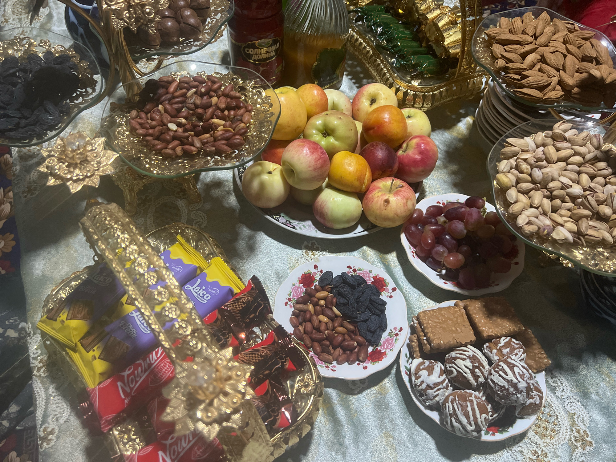 A table is laden with a festive spread of food and drinks, suggesting a celebration. The items are arranged on various platters and dishes, primarily made of glass and gold-colored metal. In the foreground, a gold ornate basket holds an assortment of chocolates wrapped in colorful foil, predominantly yellow, purple, and red. To the right of the basket, a small plate features a mix of peanuts and dark raisins, adorned with a floral pattern on its edge. Further to the right, another plate contains cookies, some square-shaped and topped with sesame seeds, and others round, chocolate-colored cookies drizzled with white icing.
In the middle ground, a collection of apples in various shades of red and yellow are piled on a white plate. To the left of the apples, a glass bowl holds a generous portion of peanuts. Behind the apples, two bottles are visible: one red bottle with Cyrillic writing that reads "СОЛНЕЧНАЯ ДОЛИНА" (Solnechnaya Dolina, meaning "Sunny Valley"), and a clear bottle containing a light-colored liquid, possibly juice or water. Above these, a tiered stand holds dark brown nuts, likely macadamia nuts or chestnuts, on its top level.
In the background, to the right of the tiered stand, a gold-colored tray contains rectangular green-wrapped items, likely candies or sweets. Two glass bowls filled with nuts are prominent on the right side of the table: one holds a large quantity of almonds, and the other is filled with pistachios. Below the pistachios, a cluster of red grapes is arranged on a plate. The table is covered with a tablecloth that has a shimmering, patterned texture in shades of blue and gray. The lighting suggests an indoor setting, possibly illuminated by overhead lights. No people or animals are visible in the frame.