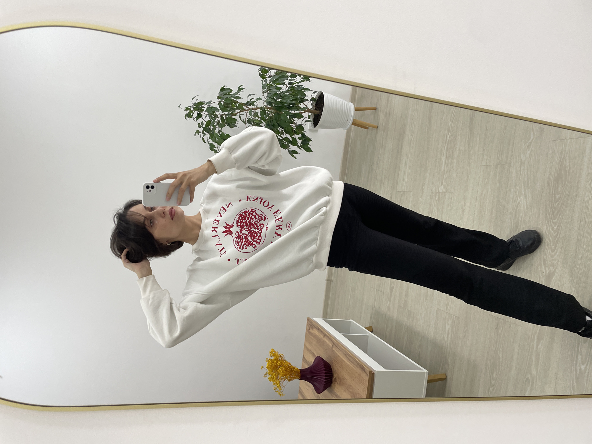 An individual with short, dark hair is captured in a full-length mirror selfie, holding a white smartphone to take the photo. The person, positioned slightly off-center to the left, is wearing a white long-sleeved sweatshirt with a red graphic design and black pants. Their right hand holds the phone, while their left hand is raised to touch their hair.
The red graphic on the sweatshirt, visible in reverse due to the mirror, features a stylized pomegranate design. Partial reversed text is visible, including what appears to be "VERITA," "ENJOY," "LOVE," and "VENEZIA," suggesting an Italian theme.
The setting, likely an indoor room in Izhevsk, Russia, is reflected in the mirror. The background consists of a plain white wall and light-colored wooden floorboards. A modern aesthetic is conveyed by the simple decor. To the right, a potted plant with lush green leaves sits in a white, ribbed pot on a light wooden stand. Below the plant, a small wooden and white storage unit with open compartments is visible. On this unit, a dark purple vase holds a small bouquet of yellow dried flowers. The person is wearing black sneakers, completing the casual outfit. The overall mood is calm and personal, as implied by the selfie activity in a tidy, minimalist space.