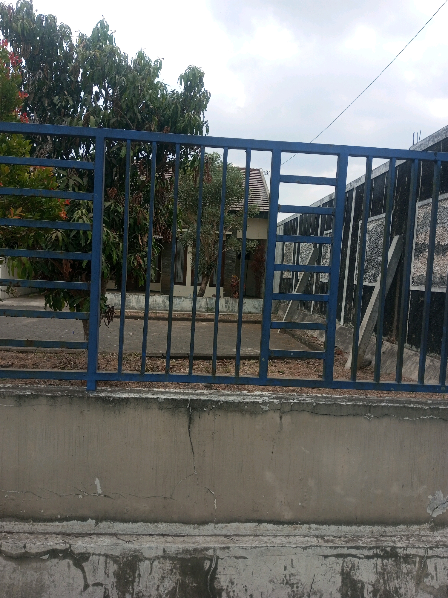 The image depicts a residential property in Pakis, Indonesia, viewed through a blue metal fence. The fence is constructed from vertical bars and horizontal beams, with a decorative pattern of square openings in its upper section. Behind the fence, a house with a tiled roof and dark-framed windows is partially visible, nestled amongst lush green trees and some drier vegetation. The ground within the property appears to be paved with interlocking bricks. To the right of the house, a dark-colored wall with textured panels rises, partially obscured by the fence. The foreground consists of a weathered concrete wall or barrier, showing signs of age and cracking. The sky above is overcast with a thin, dark electrical wire stretching across it. The scene is static, with no individuals or ongoing activities visible.