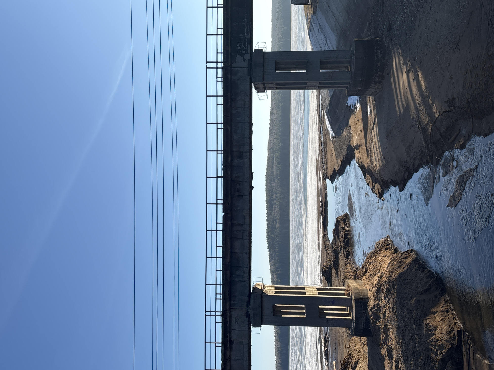 The image depicts a bridge over a body of water, likely a river or a wide stream. The bridge structure is concrete and includes support pillars. Power lines are visible against a clear blue sky. The riverbank is sandy and eroded, with water flowing between the eroded sections. The image is rotated 90 degrees counter-clockwise. While the location is presumed to be near Chaikovsky, Russia, the image itself doesn't provide definitive proof.