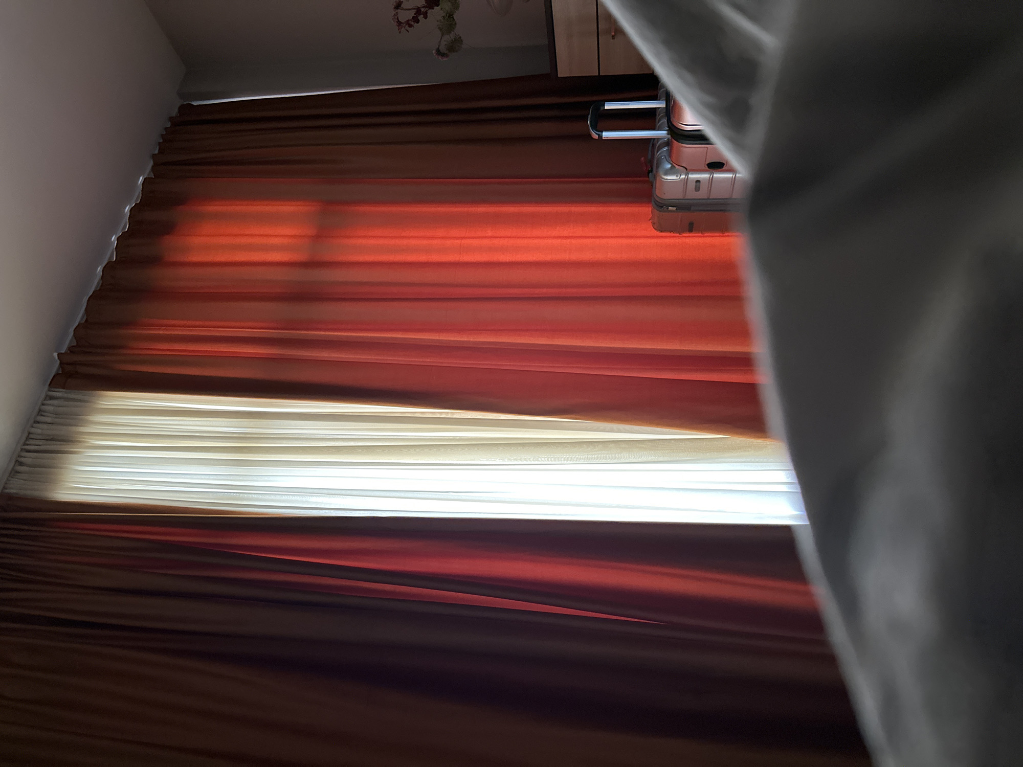 The image shows the interior of a room in Tbilisi, Georgia, with horizontal shades that are partially open, revealing bright light. A piece of luggage is visible on top of the shades. A section of fabric is partially obscuring the right side of the view. The shades have alternating brown and white sections.