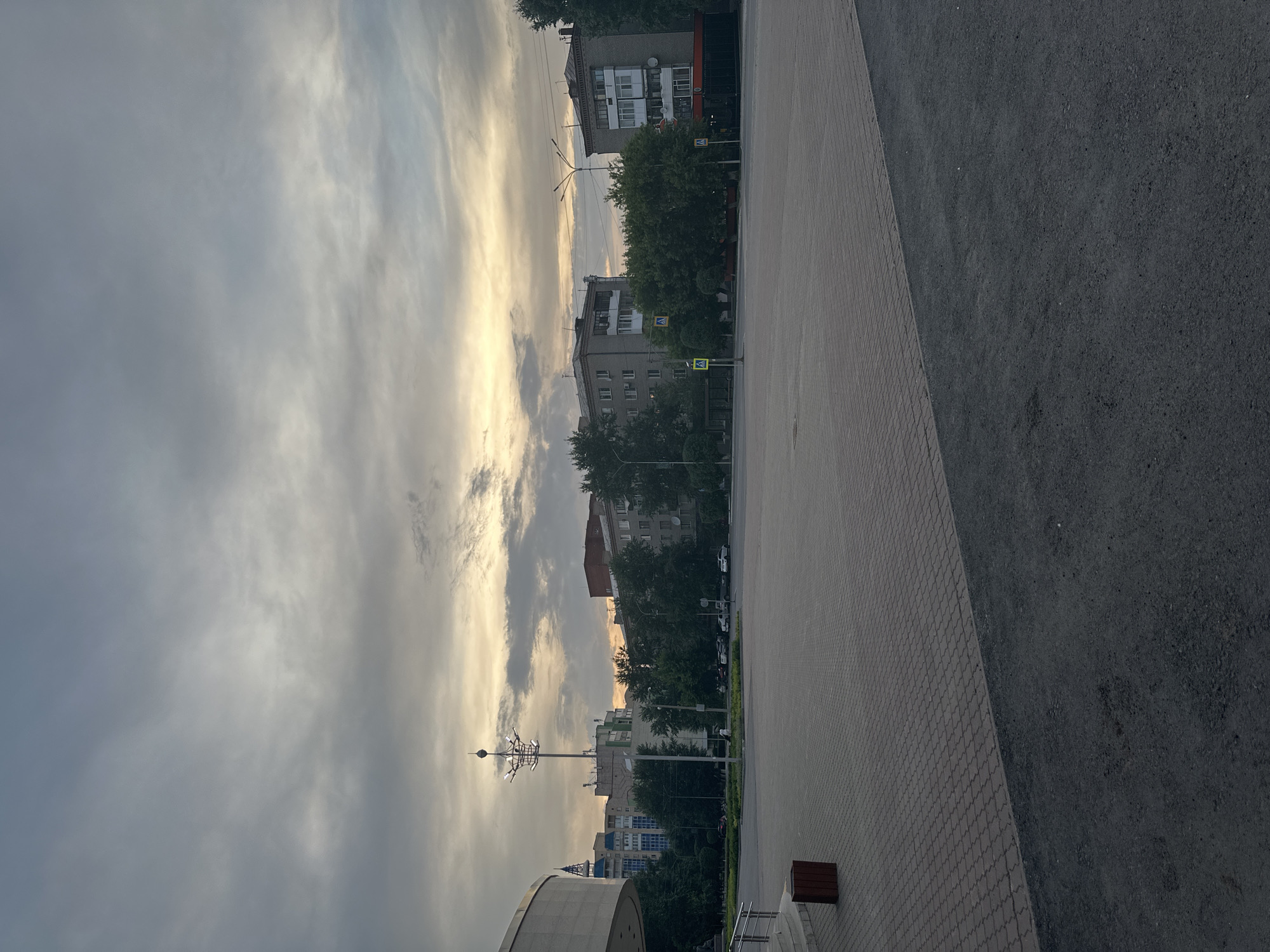 The image depicts an urban landscape under a cloudy sky with a soft, diffused light suggesting either dawn or dusk. In the foreground, a large paved area, likely a public square or plaza, is visible. The paving is composed of rectangular blocks laid in a patterned fashion. To the left, a portion of a modern, curved building with a light-colored facade is visible, along with a dark-colored trash receptacle.
In the mid-ground, a wide street separates the plaza from a line of trees and buildings. Several cars are parked along the street. A prominent street lamp with an elaborate, crown-like structure at its apex stands near the center of the scene, illuminated. Behind the trees, several apartment buildings of varying heights and architectural styles are present. Some buildings are light-colored, while others are darker brick. Pedestrian crosswalk signs are visible on the street.
The background is dominated by the sky, which is filled with a complex pattern of grey and white clouds, with a hint of yellow or orange light breaking through in the distance, indicating the direction of the sun. The overall impression is of a quiet, possibly residential or civic area in a city. Based on the visual cues, such as the architectural style of the buildings and the presence of specific street furniture, this scene is identifiable as Kokshetau, Kazakhstan, likely in the vicinity of the city's cultural or administrative center. No people are clearly visible in the image.