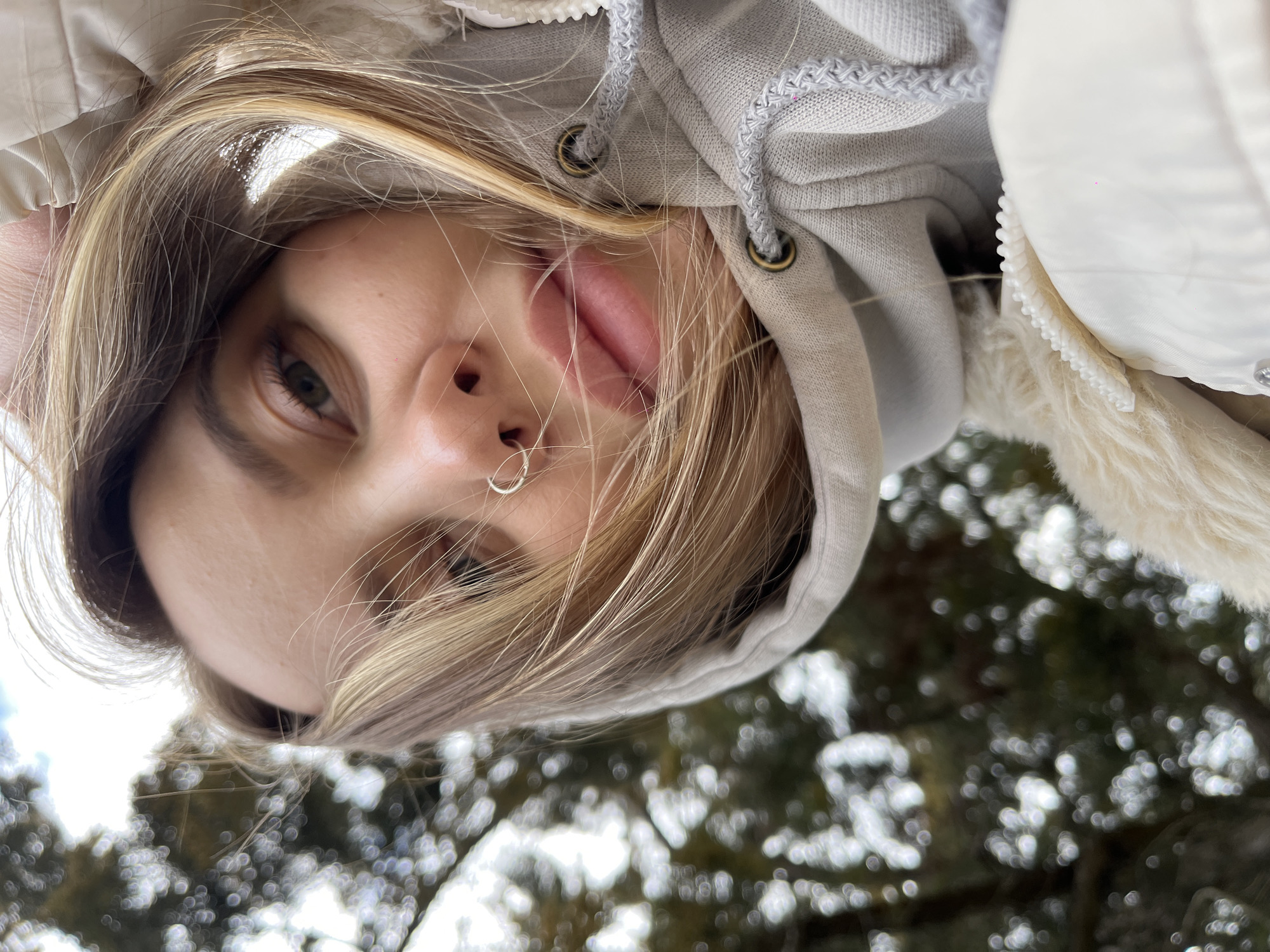 A young woman with blonde hair and a nose ring is holding a phone and taking a selfie. She is outdoors and wearing a grey hooded sweatshirt with a white fuzzy lined coat. She is sticking her tongue out. The trees and sky are visible in the background. The picture was taken in Novopokrovskaya, Russia.