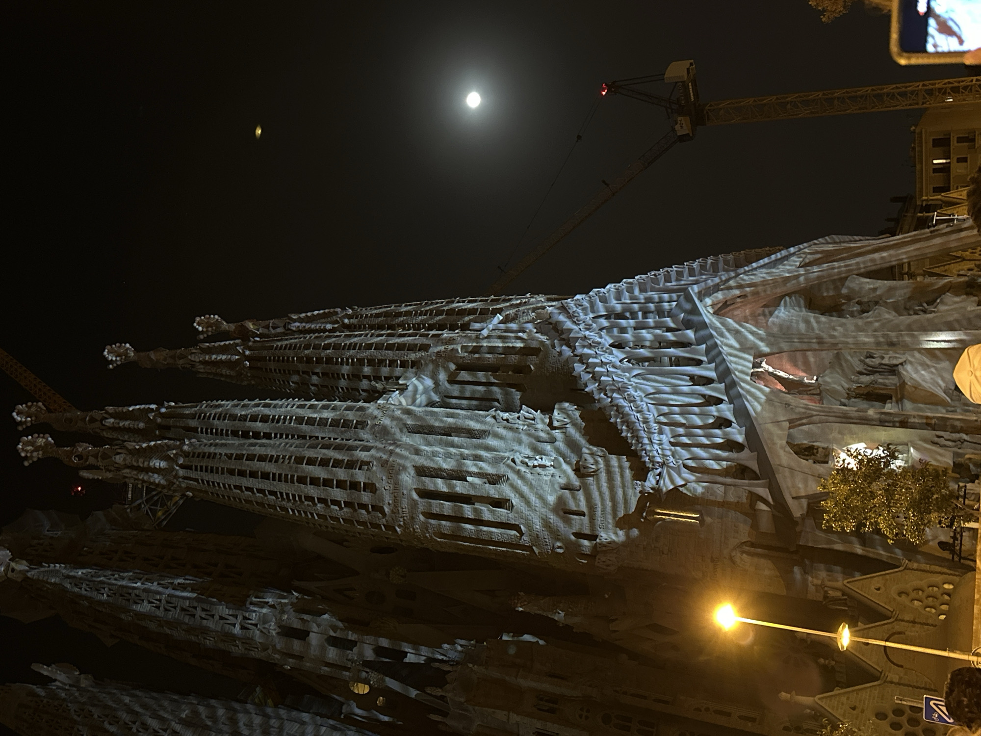 The image shows the Sagrada Familia basilica in Barcelona, Spain, at night. The structure is the main subject, with its intricate spires and facade illuminated. A construction crane is visible in the background, indicating ongoing work. A street lamp shines brightly in the lower right, and the moon is prominent in the upper portion of the frame. A building is behind Sagrada Familia. People stand in the right corner of the frame.