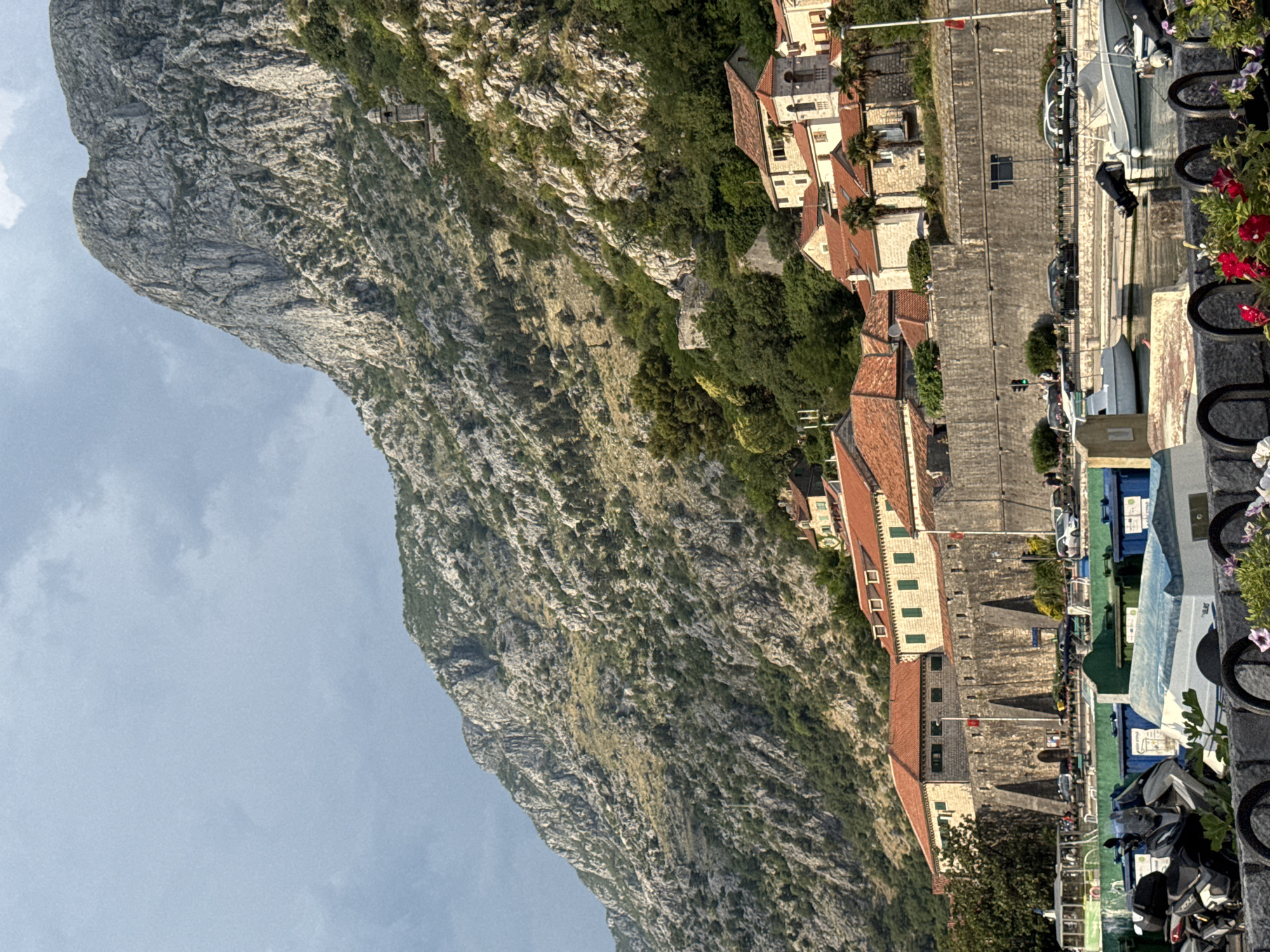 The image captures a scenic view of the city of Kotor, Montenegro, with its historic fortifications and the imposing mountains that surround it. In the foreground, a marina is visible with several boats docked, including a white motorboat with a black outboard engine and another white boat with a blue tarp. Some trash bins are also present in the foreground, with a motorcycle parked to the left. A stone wall, part of the city's fortifications, runs horizontally across the mid-ground, with buildings featuring red tile roofs situated behind it. These buildings have light-colored facades and green shutters. The steep, rocky mountain slopes, covered in sparse vegetation, dominate the background, with a church tower visible high up on the mountainside. The sky is overcast with dramatic clouds.