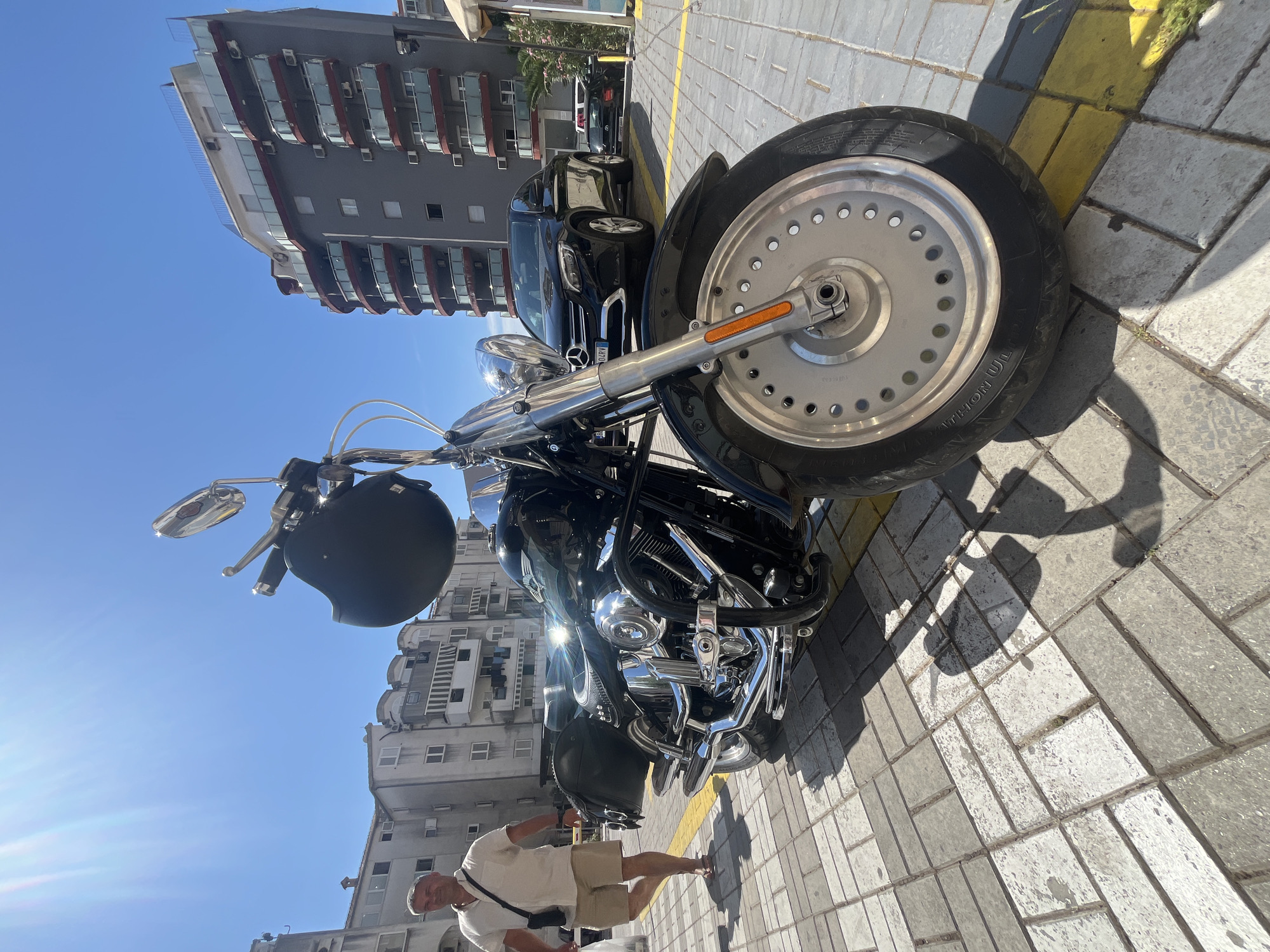 The image, captured in Donji Pobori, Montenegro, presents a bright, sunny street scene, rotated 90 degrees counter-clockwise from its natural orientation. The time of day appears to be midday or early afternoon, indicated by the clear blue sky and strong shadows cast by objects.
The central subject is a large, black cruiser-style motorcycle, possibly a Harley-Davidson, parked on a paved area. It features prominent chrome accents on its engine, exhaust pipes, and handlebars. A black motorcycle helmet hangs from the left handlebar. The motorcycle has a distinctive solid, disc-like front wheel, with the text "MARATHON UL" partially visible on its tire sidewall.
Standing next to the motorcycle, closer to the viewer, is a man. He wears a light-colored, short-sleeved collared shirt, light-colored shorts, and sandals, with a black cross-body bag slung over his shoulder. He is looking towards the camera.
In the background, several multi-story buildings line the street. One building is grey with dark reddish-brown curved balconies, while another is a lighter grey or beige. Behind the motorcycle, a dark-colored SUV, which appears to be a Mercedes-Benz, is parked. Its license plate is partially visible, showing "04RVX". Another vehicle is indistinctly visible further back.
The ground is paved with light grey and off-white rectangular blocks, resembling cobblestones or pavers. Yellow lines, likely indicating parking spaces or road markings, are visible on the ground. The overall setting is an urban or semi-urban environment on a clear, warm day.