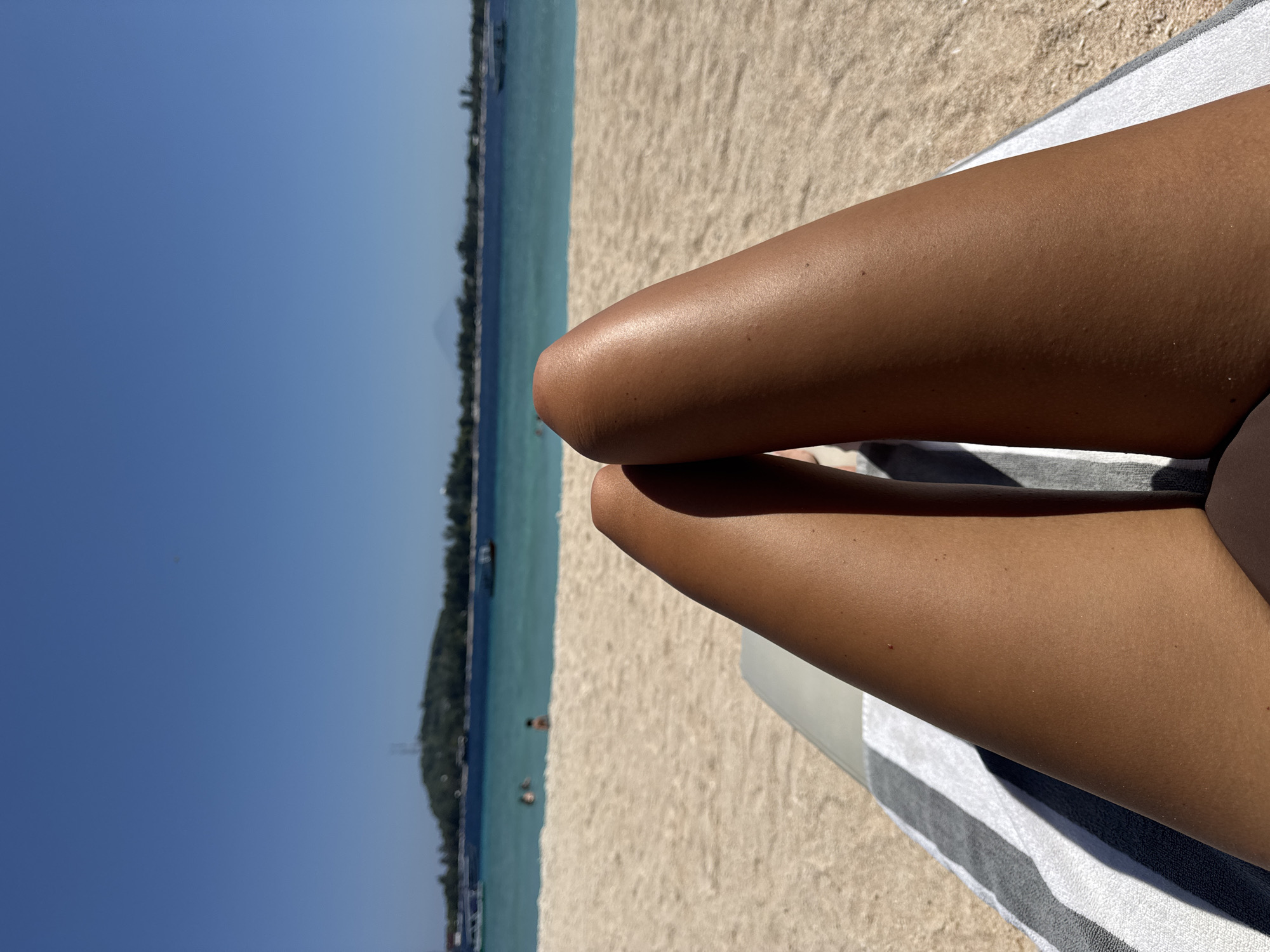 The image shows a pair of crossed legs on a beach, presumably lying on a towel with grey and white stripes. The background shows a beach with light-colored sand and a body of water with a tree-lined shore in the distance. The water is blue-green and the sky is clear blue. Some people are visible in the water. The location is Unknown, Indonesia.
