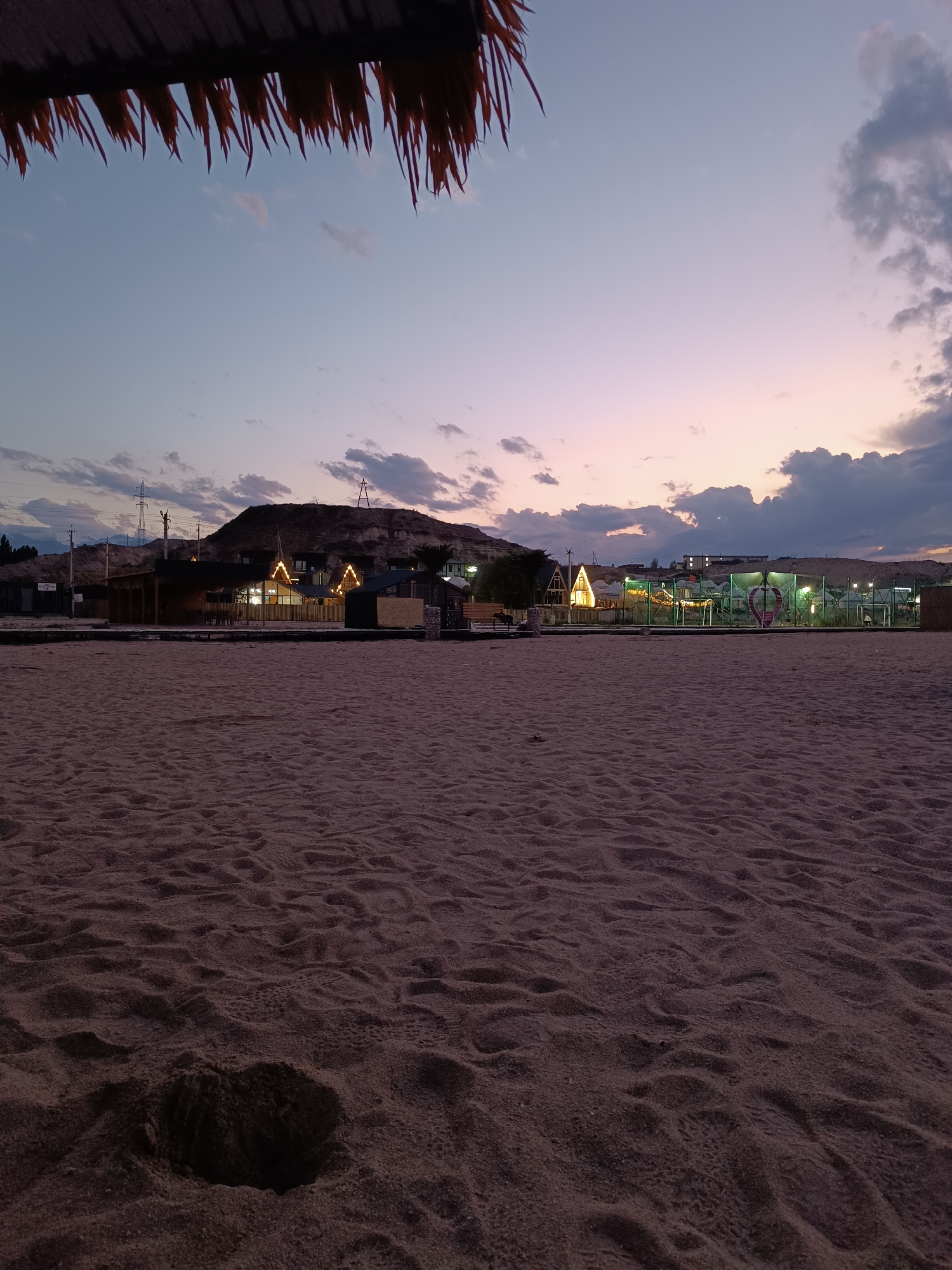 The image depicts a tranquil beach scene at dusk, with the sky transitioning through soft hues of blue, purple, and pink. In the foreground, the sand is textured with ripples and a small, dark indentation, possibly a sand crab hole or a footprint. A thatched roof overhangs the top left of the frame, casting a shadow. In the mid-ground, a row of small, illuminated cabins with triangular roofs are visible, suggesting a resort or a cluster of holiday homes. These structures are nestled against a backdrop of a barren, hilly landscape. Power lines and pylons are also visible in the distance, indicating proximity to a populated area. The overall atmosphere is peaceful and serene, characteristic of a quiet evening by the water. The image is not specific to the city of Tong, Kyrgyzstan, as no distinct landmarks or identifying features suggest this particular location.