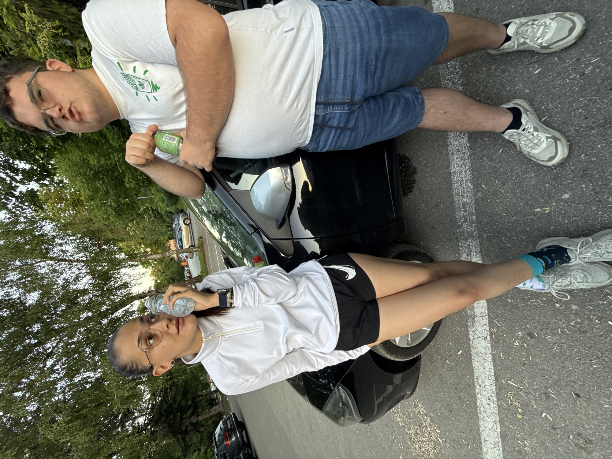 Two people pose on or near a black car parked in an outdoor parking lot, likely in a city but not identifiable as Pleven, Bulgaria from the image. A male on the upper right wears a white t-shirt with a green design, denim shorts, and gray sneakers with dark socks. He is holding a green can. A female on the lower left wears a white long sleeve, black athletic shorts, gray sneakers, and patterned blue and green socks. She holds a water bottle. Both people are wearing glasses. The parking lot is paved with a white line separating parking spaces. Green trees are visible in the background.