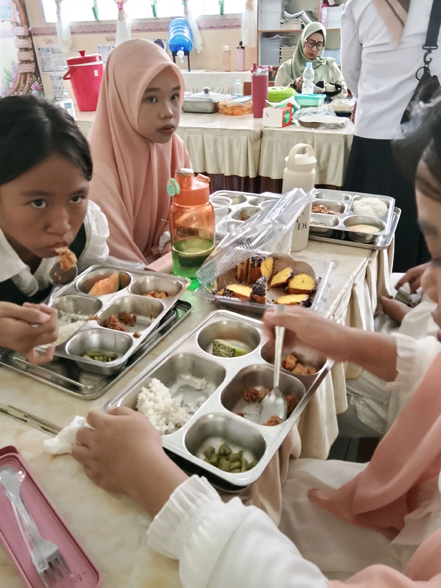 In the city of Besusu Timur, Indonesia, a group of young girls are seated at a table, engaged in a meal. The table is covered with a light-colored tablecloth, and on it are several metal trays containing food. In the foreground, a girl wearing a white shirt with ruffled sleeves is actively eating from a tray with a spoon. Her tray contains rice, green beans, and other unidentified food items. To her right, another girl, partially visible, is also attending to her meal. In the middle ground, a girl wearing a pink hijab and a pink headscarf is looking towards the camera, with her food tray in front of her. Next to her is an orange water bottle with a green lid. Further in the background, a woman in a green hijab and glasses is seated at a table, interacting with her food. The environment appears to be an indoor setting, possibly a school or community hall, with shelves and a water cooler visible in the background. The lighting suggests it is daytime.