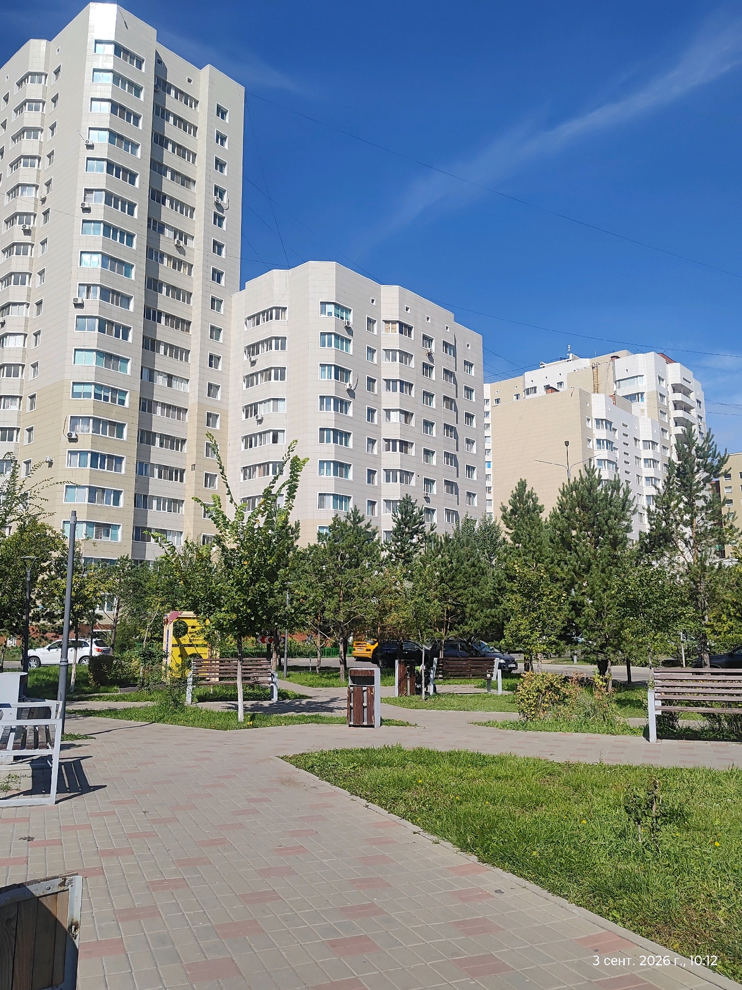 The image depicts a residential area in Astana, Kazakhstan, on a clear day. The foreground features a paved walkway with a pattern of gray and reddish-brown square tiles, leading into a grassy area with scattered yellow flowers. Several park benches are visible, some made of wood and metal, others appearing to be white metal structures. A brown trash receptacle is situated near one of the benches.
In the middle ground, a cluster of trees, including both deciduous and coniferous varieties, creates a natural border. Behind the trees, several modern, multi-story apartment buildings dominate the scene. The buildings are primarily light-colored, with numerous windows and balconies. One prominent building on the left is tall and octagonal in shape. To the right, other apartment buildings of varying heights and architectural styles are visible, including one with a light beige facade. A yellow structure, possibly a public utility box or a decorative element, is located among the trees.
Several vehicles are present, including a white SUV parked on the left side of the walkway, a dark-colored car partially obscured by trees, and a yellow vehicle further back. Power lines are strung across the bright blue sky, with a few wispy clouds. A timestamp in the bottom right corner reads "3 сент. 2026 г., 10:12". No people are clearly visible in the image.