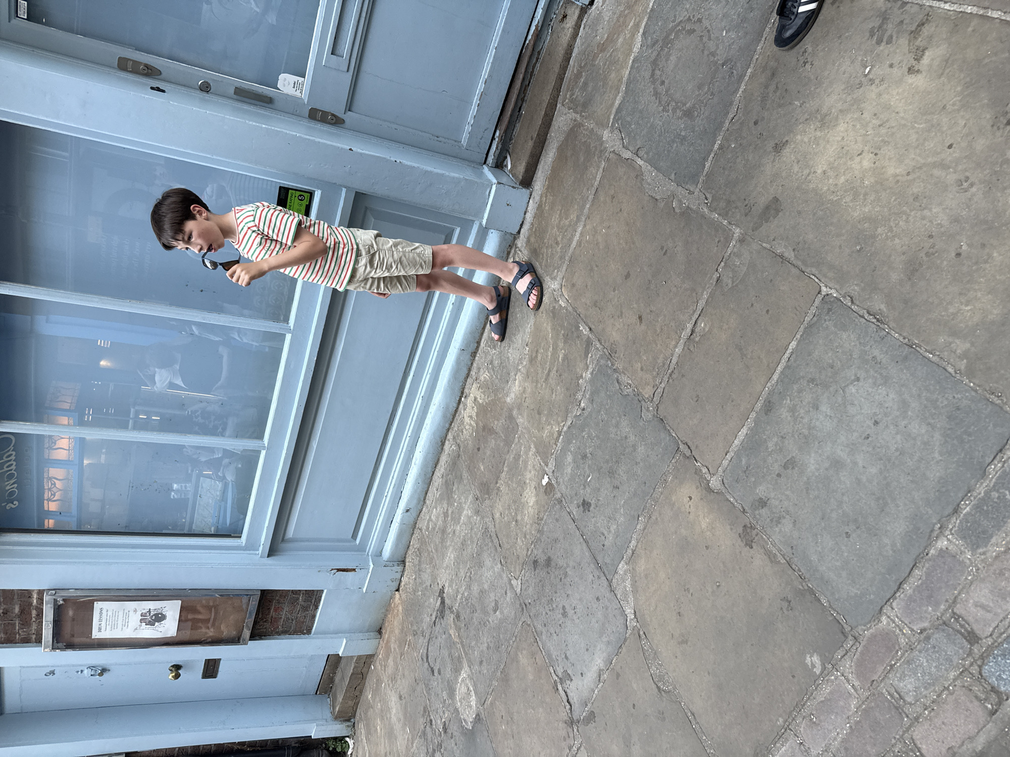 A young boy stands on a stone-paved street in front of a light blue building facade, possibly in London, United Kingdom. He is wearing a striped shirt, khaki shorts, and sandals, and appears to be examining something through a pair of eyeglasses he holds up to his face. The building has a large window and a lower door with a sign displayed. Part of a shoe is visible in the upper right corner of the image. The scene is captured in daylight, with the angle of the photograph slightly tilted to the right.