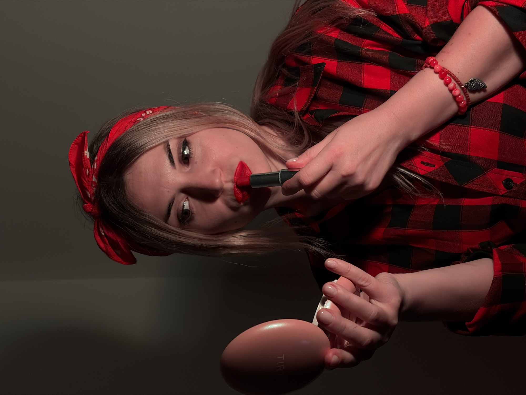 A woman applies red lipstick while holding a pink compact. She wears a red and black plaid shirt and a red bandana. The background is a dark gray wall. It is not possible to confirm the location is in Budva, Montenegro from the image.