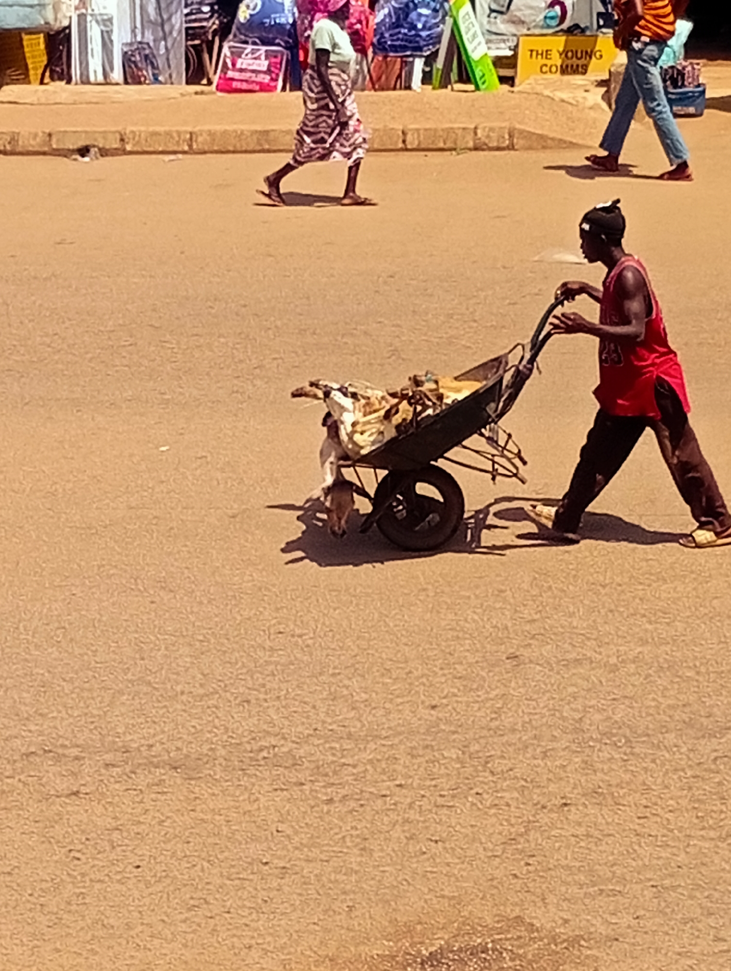 In Bukuru, Nigeria, a young man wearing a red sleeveless shirt, black pants, and yellow sandals pushes a wheelbarrow laden with what appears to be refuse or discarded materials along a dusty, sun-baked road. He is partially obscured by the wheelbarrow, with his face turned to the left. In the background, a woman in a patterned dress walks away from the camera, and other people and market stalls are visible. The scene is illuminated by bright sunlight, casting distinct shadows on the ground.