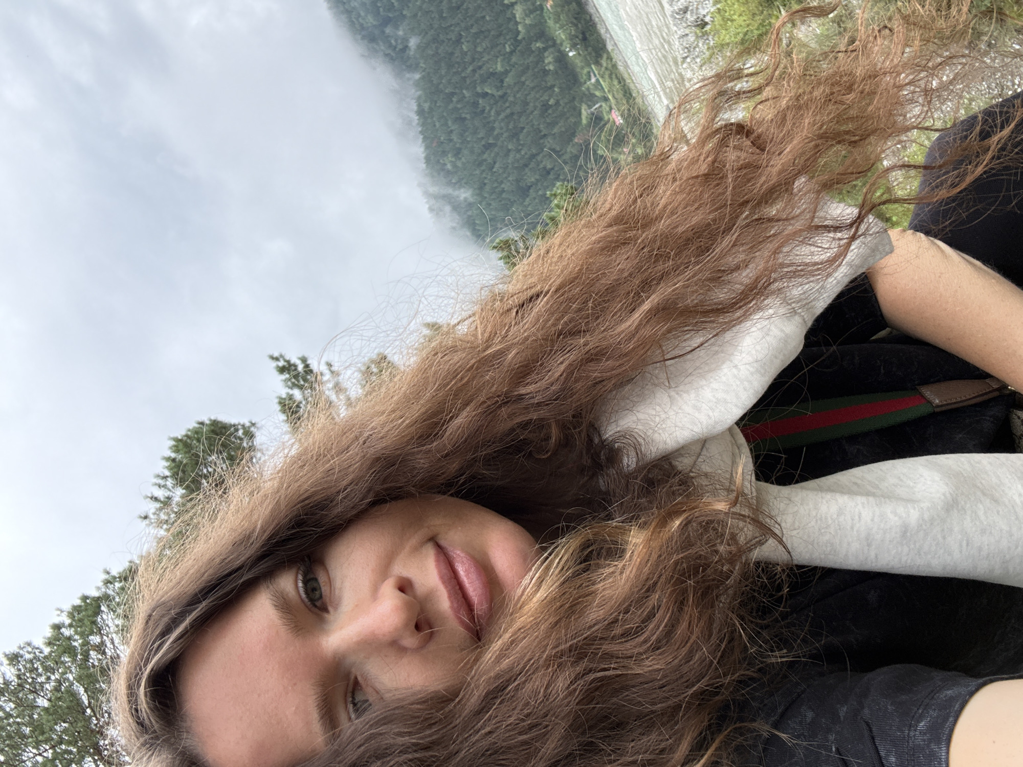 In a selfie taken outdoors, a woman with long, wavy brown hair is in the foreground, with her face angled towards the camera. She has fair skin, green eyes, and is wearing a light-colored scarf over a dark top. Behind her, a lush, green, forested mountainside rises, partially obscured by mist. A body of water, likely a river, is visible in the lower right portion of the background, with rocky terrain along its edge. The sky above is overcast and cloudy. The woman appears to be in a natural, mountainous landscape, possibly near Elekmonar, Russia, given the context.