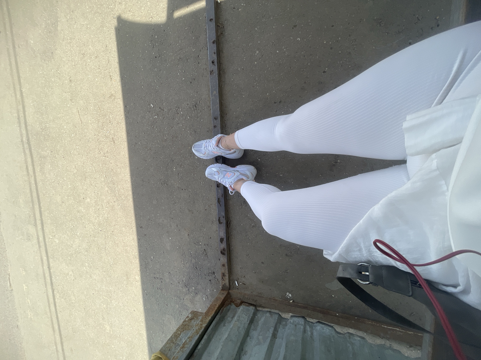 The image shows a person wearing white ribbed leggings and light grey and pink sneakers. They are standing on a concrete surface next to a rusty metal structure with holes, possibly a drain or part of a building. The person is also wearing a white garment, and a black bag with a red strap is visible. The specific location in the city of Molokovo, Russia, cannot be determined from the image.