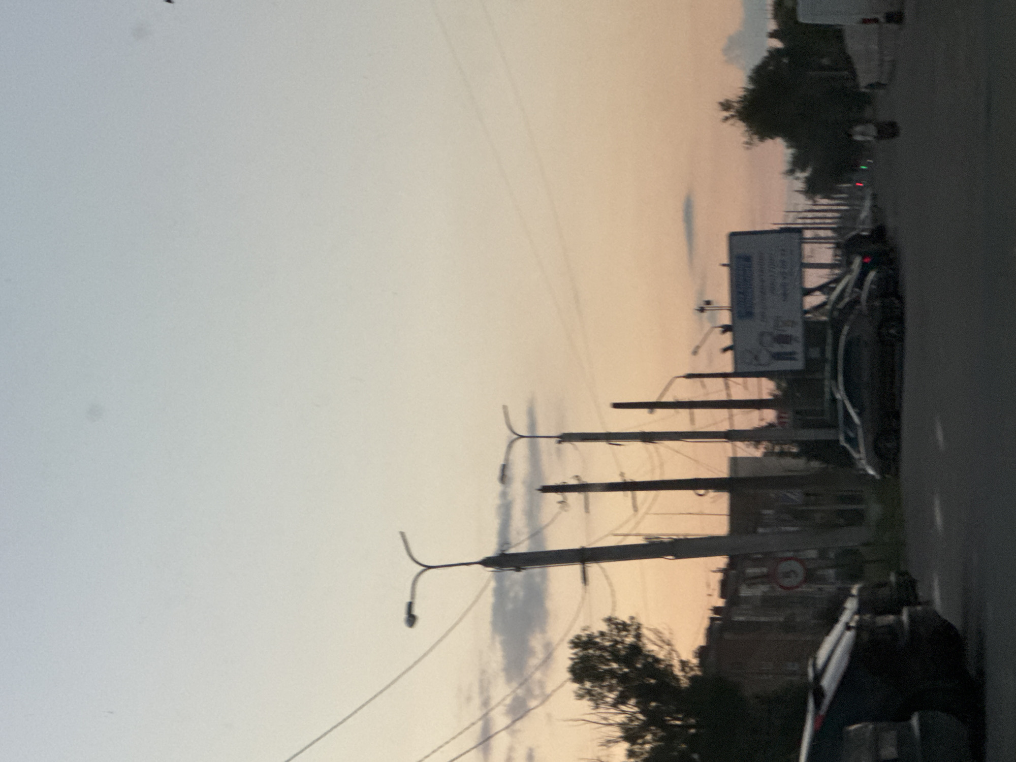 In this photograph, a street scene unfolds at dusk in an urban setting. The sky displays a gradient of soft, warm hues, transitioning from a pale blue at the top to a peachy orange near the horizon, with faint clouds present. Power lines and streetlights dominate the mid-ground and background, with several tall poles supporting multiple lights and cables stretching across the frame.
On the left side of the image, the dark silhouettes of trees are visible against the colorful sky, adjacent to a row of multi-story buildings. A traffic sign, circular with a red border and a horizontal line, is mounted on a pole.
The foreground is occupied by parked cars. To the left, a dark-colored sedan is partially visible, facing away from the viewer. Further down the street, a line of other cars is parked. In the distance, a large billboard is mounted on poles, displaying text and an illustration. A solitary figure walks in the distance. The overall impression is one of a quiet street at the end of the day, with artificial lighting beginning to illuminate the scene.