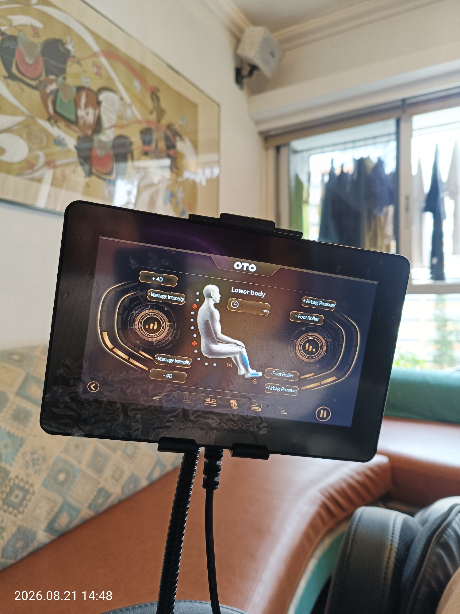 A tablet screen displays a massage chair control interface, showing options for "Lower body" massage, with controls for "Massage Intensity," "+4D," "-4D," "Airbag Pressure," and "Foot Roller." A silhouette of a person indicates the targeted massage area. The tablet is mounted on a flexible arm, suggesting it's part of a massage chair. In the background, a portion of a room is visible, including a painting on the wall, a window with laundry hanging outside, and parts of a brown leather couch. The timestamp "2026.08.21 14:48" is visible in the lower-left corner of the image, suggesting this is a contemporary scene. The overall setting appears to be an indoor residential or commercial space. The specific location in Hong Kong, China cannot be determined from the visual information alone.