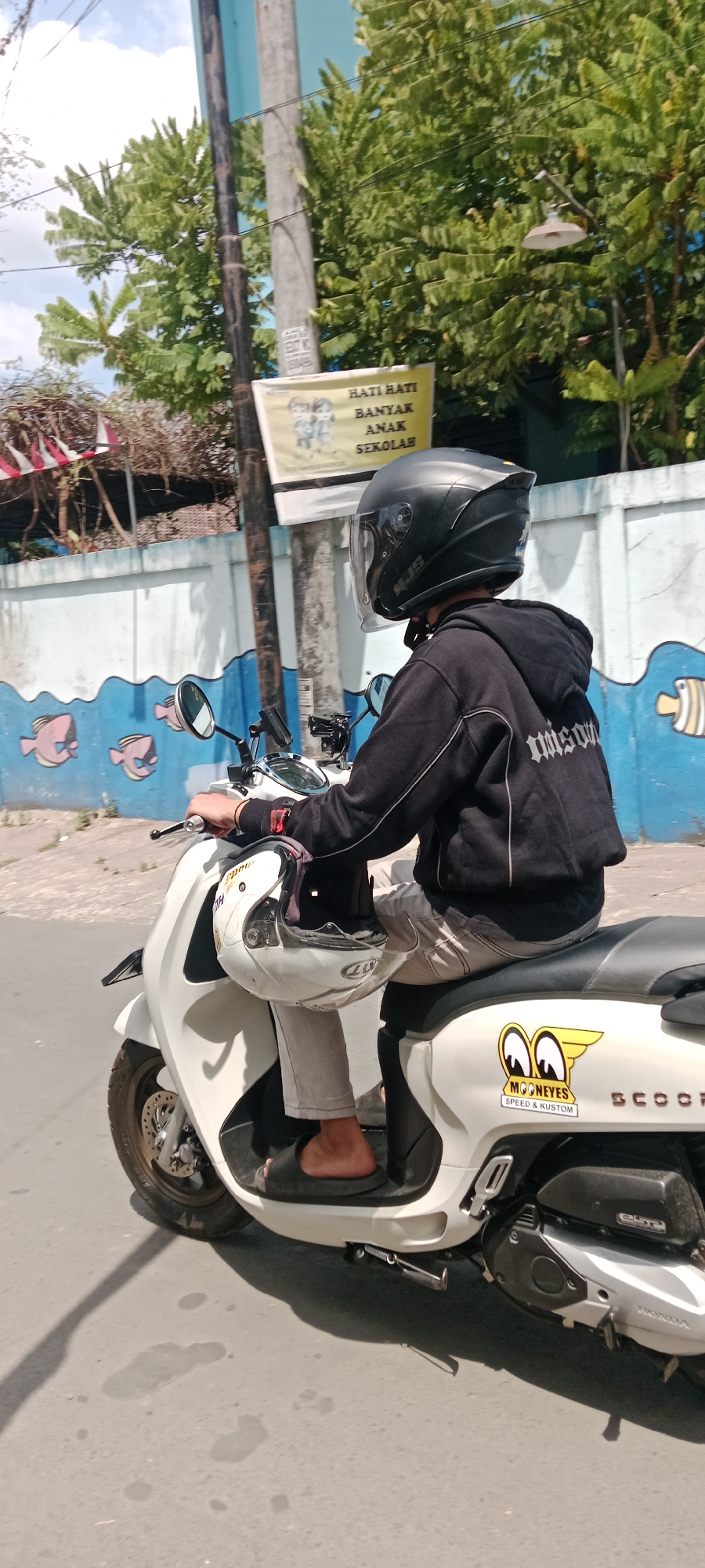 A person wearing a black helmet and a black hoodie is riding a white scooter. The scooter has a "MOONEYES SPEED & KUSTUM" sticker on the side. The person is holding a white helmet in their left hand. The scooter is on a street in Purwomartani, Indonesia, with a blue and white wall with fish drawings in the background. There are trees and a cloudy sky visible. A sign with Indonesian text "HATI HATI BANYAK ANAK SEKOLAH" which translates to "BE CAREFUL, MANY SCHOOL CHILDREN" is also present.
