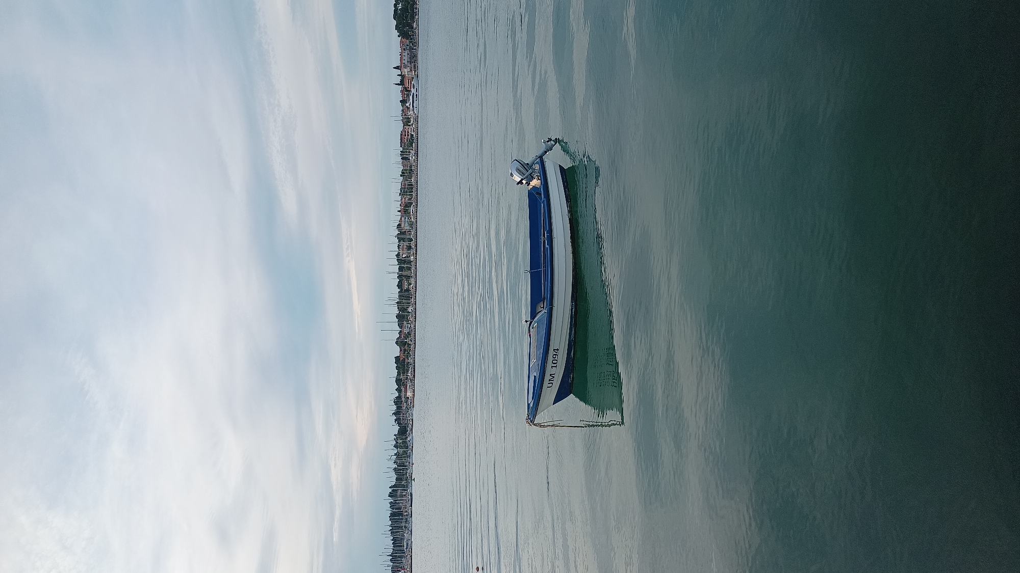 The image displays a tranquil coastal scene, featuring a single motorboat floating on calm, reflective water. The photograph, when viewed correctly, is horizontally oriented, presenting a wide expanse of water in the foreground and midground, a distant coastline, and a bright sky overhead.
The primary subject is a small, blue and white boat equipped with an outboard motor at its stern. The registration number "UM 1094" is clearly visible on the side of its hull. The boat appears to be stationary, possibly anchored or moored, and its distinct reflection shimmers gently on the water's surface. The water itself is a clear, greenish-blue hue, with slight ripples indicating a gentle movement.
In the background, a long, low-lying coastline stretches across the horizon. This shoreline is dotted with buildings, suggesting a town or settlement, and numerous masts indicate the presence of a harbor or marina with many docked boats. Green foliage is also visible along the coast.
The sky above is predominantly light blue and clear, with sparse, wispy white clouds adding texture without obscuring the view. The soft, even lighting suggests either an early morning or late afternoon setting, contributing to the overall peaceful ambiance. There are no people visible in the scene. Given the location context of Umag, Croatia, the scene likely captures a quiet moment in the Adriatic Sea.