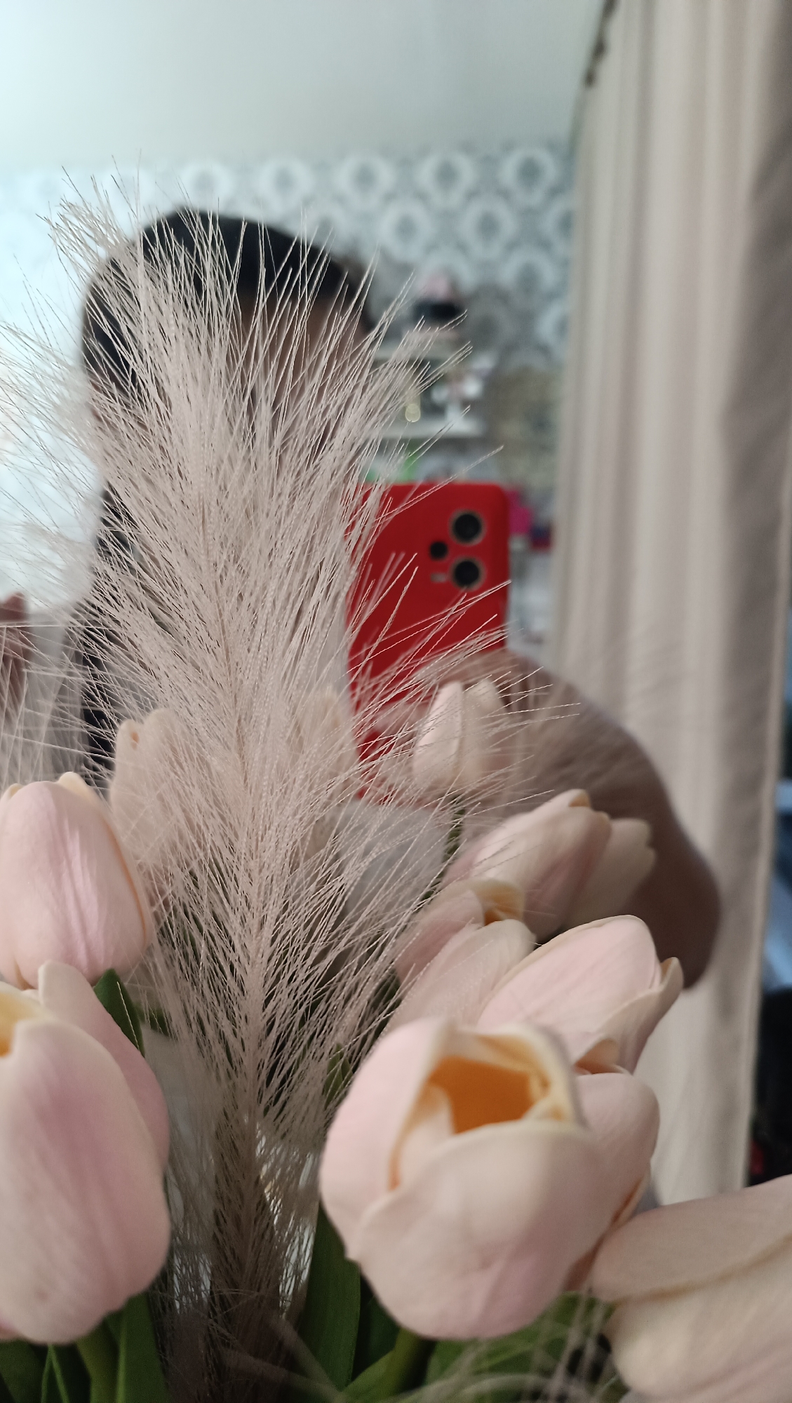 The image shows a close-up of a bouquet of light pink tulips, with a feathery ornamental grass stem in the foreground. In the blurred background, a red smartphone is held up to take a picture, reflecting a person's face and hair. The overall scene is indoors, with hints of a room's decor including a patterned wallpaper and curtains. There is no information in the image to suggest a location in Chaikovsky, Russia.