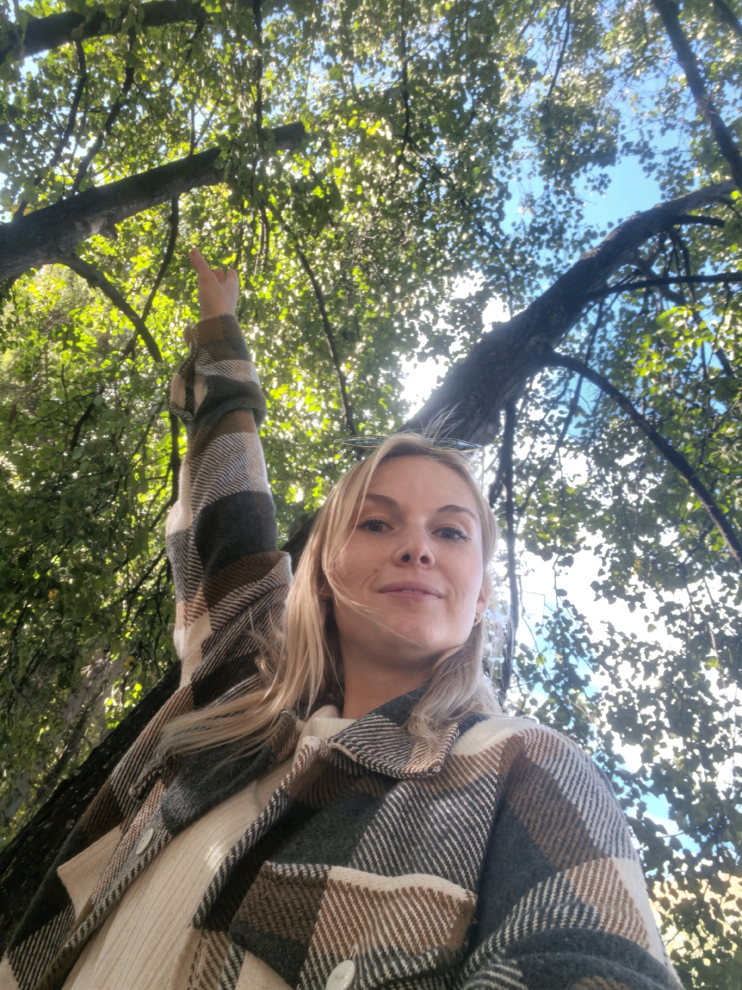 A young woman is pictured from a low angle, looking up towards the camera with a slight smile. She has blonde hair and is wearing a cream-colored sweater under a plaid jacket with brown, grey, and white patterns. Her left arm is raised, pointing upwards towards the tree branches and the bright blue sky visible through the foliage. The setting appears to be outdoors, under a canopy of green leaves and tree limbs. No specific location in Sterlitamak, Russia can be determined from the image alone.