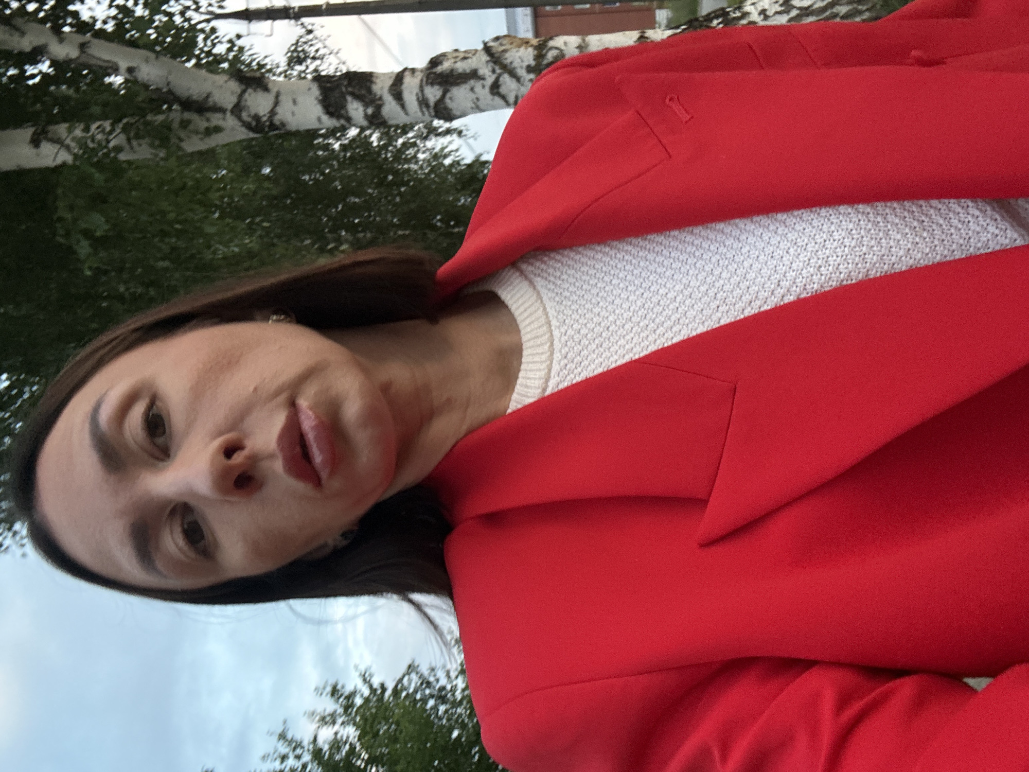 A woman with dark brown hair is taking a selfie outdoors in Votkinsk, Russia, in front of a birch tree. She is wearing a bright red blazer over a white textured sweater. The background shows a cloudy sky and green foliage. The image is framed from the shoulders up, with the woman's face centrally located. The lighting suggests it is daytime, possibly late afternoon or early evening.