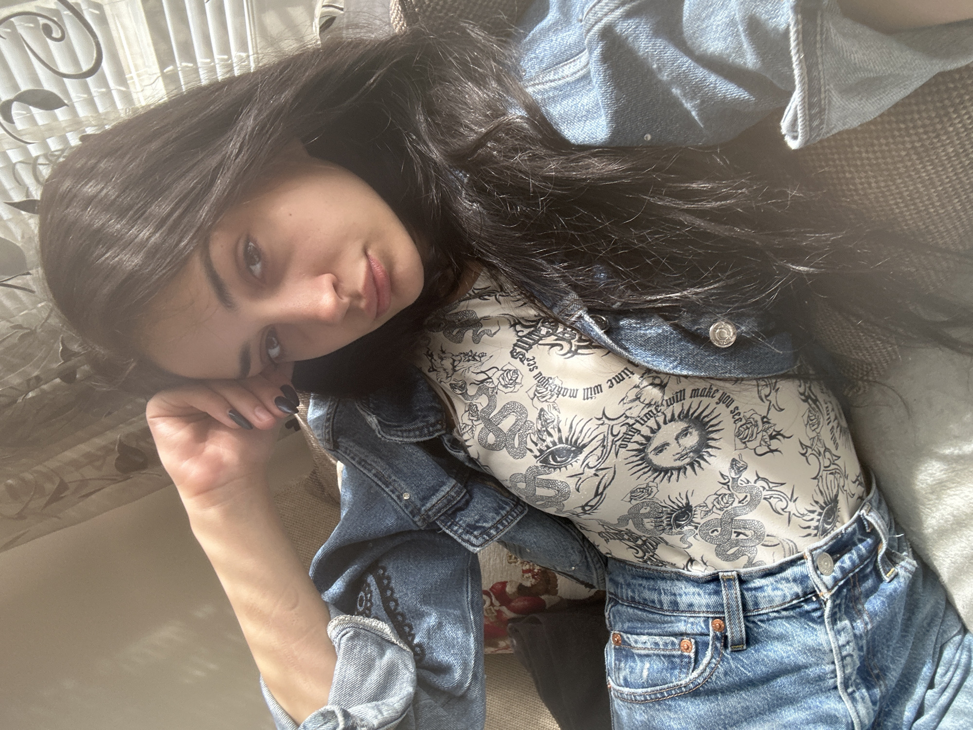 A young woman is lying down, likely indoors, illuminated by bright natural light coming through window blinds. She has long, dark hair and is resting her head on her hand, looking towards the camera. She is wearing a denim jacket over a white top with black graphic prints, and denim jeans. The scene suggests a casual, relaxed setting, possibly within a home. The exact location in Burgas, Bulgaria cannot be determined from the image alone.