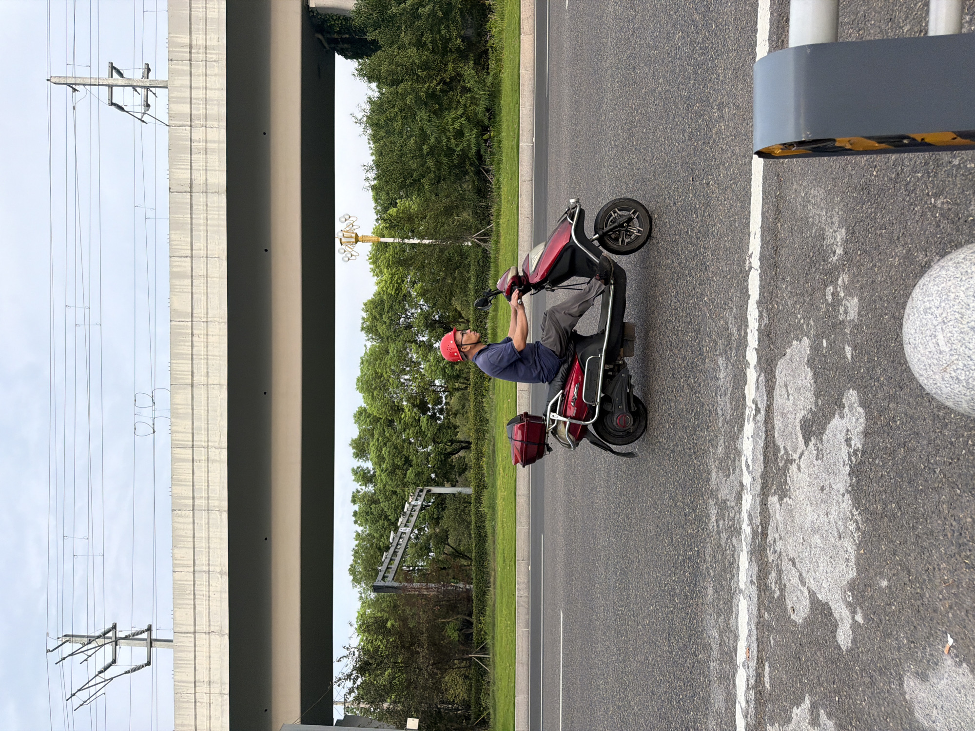 An adult male wearing a red helmet and glasses rides a red electric scooter on an asphalt road in Jiangjin, China. He is wearing a dark blue t-shirt and gray pants. The scooter has a red storage box on the back. In the background, there is a concrete overpass with overhead power lines. Green trees and vegetation line the side of the road, and a lamppost is visible in the distance. The sky is overcast.
