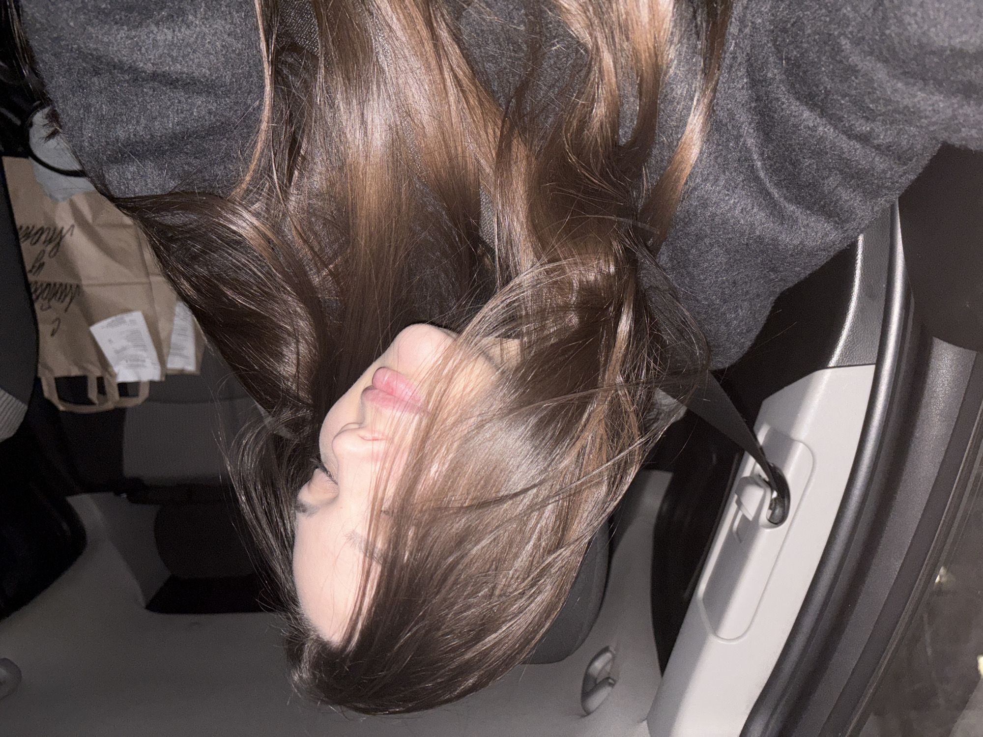A person with long brown hair is in a car. They are wearing a gray coat. A shopping bag is visible in the background. The car is stopped or parked, as the person is not actively driving. The location in Chaikovsky, Russia, is not discernible from the image.