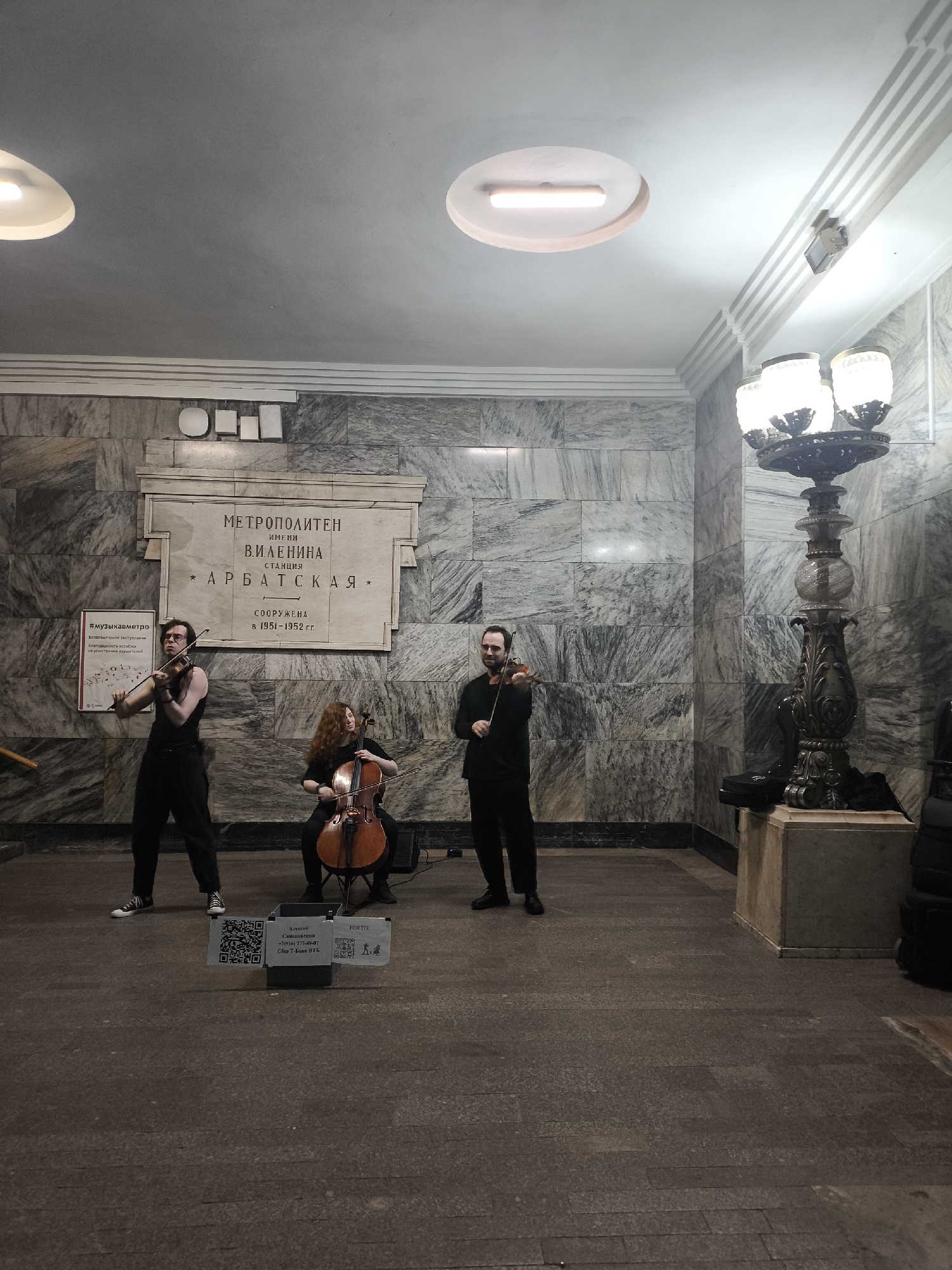A wide-angle shot reveals a three-person string ensemble performing within the Arbatkaya station of the Moscow Metro. The ensemble consists of two violinists, male and female, standing and a female cellist seated between them. The musicians are dressed in black. The ensemble stands in front of a plaque, engraved with Russian text, reading "METROPOLITEN" and "* ARBATSKAYA *". A sign with QR codes stands in front of the musicians. A vintage-style lamp post is to the right of the group. The station interior consists of gray marble.