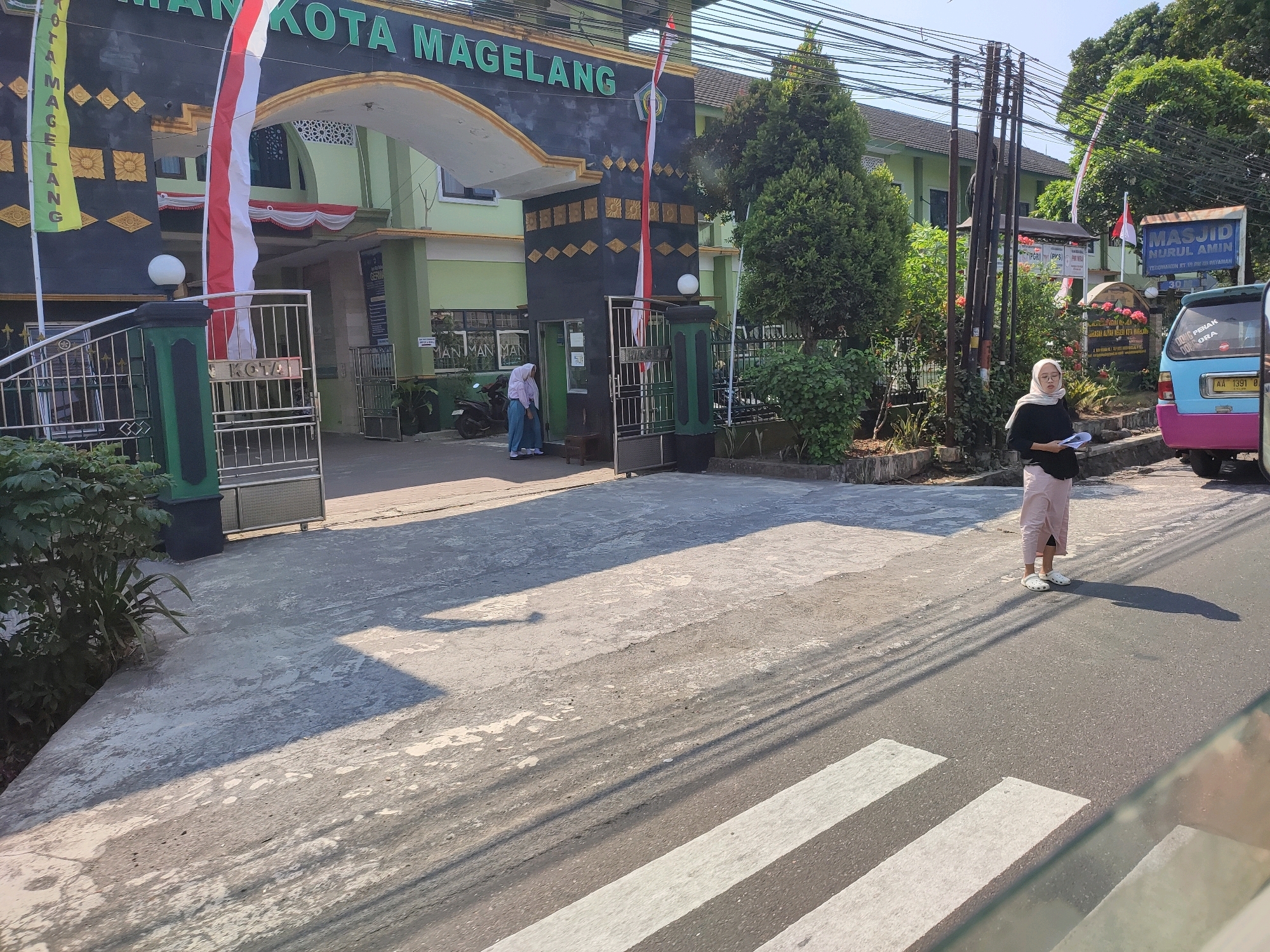 The image shows the exterior of a building with text that appears to say "KOTA MAGELANG". In front of the building is a paved area with a pedestrian crossing visible in the foreground. Two individuals are present: one person in a white hijab and pink skirt stands on the sidewalk, holding a document, and another person, also in a hijab, walks near a motorcycle by an entrance. A blue and pink vehicle is parked on the right side of the road. The environment is sunny with shadows cast on the ground. A sign for "MASJID NURUL AMIN" is visible on the right, indicating the location might be near a mosque in Payaman, Indonesia.