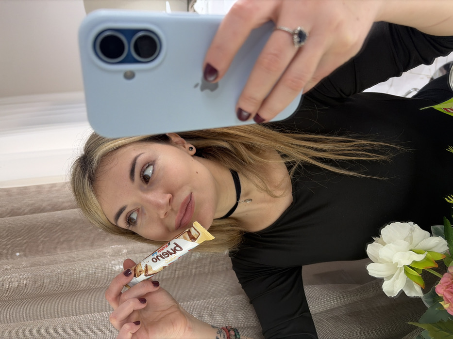 A female individual with blonde hair, wearing a black long-sleeved top, is depicted in an indoor setting, taking a mirror selfie. She holds a white-packaged Kinder Bueno White snack bar in her right hand, positioned near her mouth. Her left hand holds a light blue cased iPhone with visible dual rear cameras and an Apple logo, directed towards the viewer. Dark nail polish is applied to her fingernails, and a ring with a dark central stone surrounded by smaller stones is on her left ring finger. She also wears a black choker with a small metallic charm and small dark stud earrings. Her gaze is directed upwards and to the right. The immediate foreground includes artificial white and pink flowers. The background features a light-colored, sheer curtain on the left and a blurred, light-toned interior space on the right. The specific location within the city of Budva, Montenegro, cannot be determined from the visual information provided.
