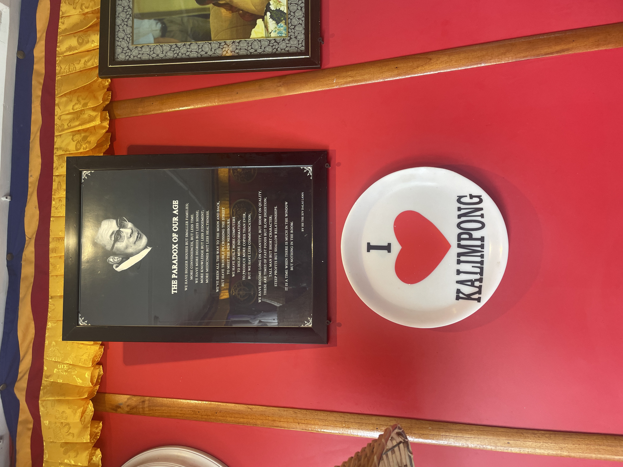The image depicts a framed photograph of the Dalai Lama mounted on a vibrant red wall. The photograph contains text titled "THE PARADOX OF OUR AGE." Below the framed photograph, a white plate with a red heart and the text "KALIMPONG" is displayed. The wall is adorned with yellow and red fabric draped along the top. To the right of the main photograph, another framed picture is visible, though its contents are partially obscured. A wooden pole runs vertically down the center of the image, and to the left, a portion of a white circular object is visible. The scene appears to be an interior setting, possibly a shop or a home, in Bhagsu, India.