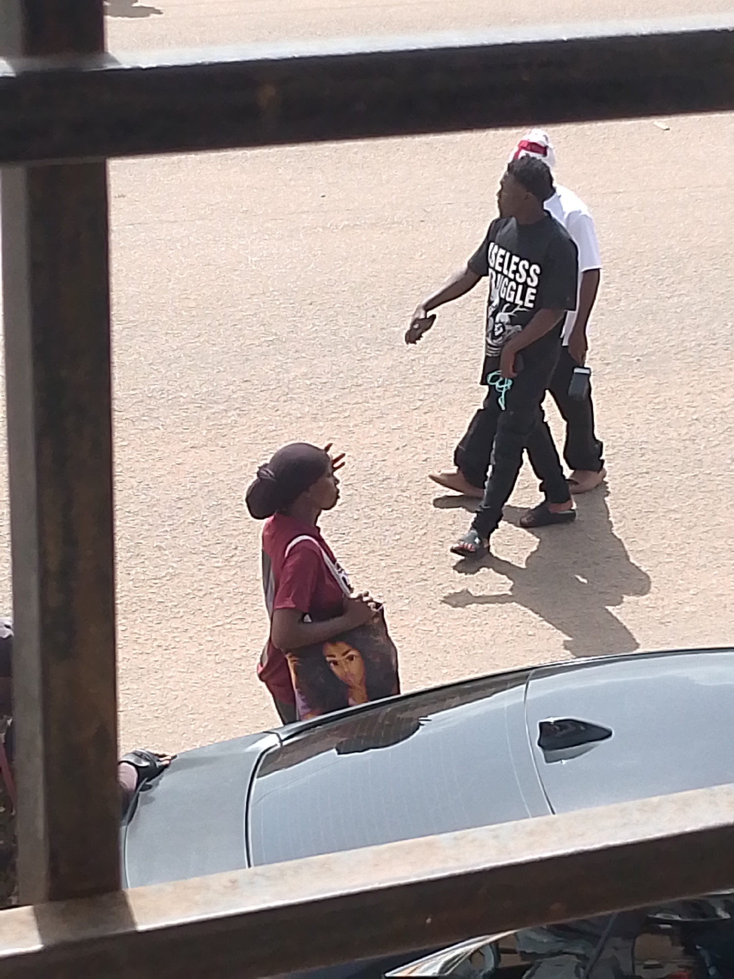 In Bukuru, Nigeria, three individuals are visible on a paved surface, possibly a street or walkway, with a dark-colored car in the foreground. A person wearing a dark t-shirt with white lettering that reads "GELESS UGGE" and dark pants is walking away from the camera. Behind this individual, another person in a white t-shirt and a red and white head covering is also walking. To the left, a person wearing a maroon t-shirt and dark pants is facing away from the camera, holding a large tote bag with a graphic of a woman's face. A dark-colored car, likely a sedan, is parked or stopped in the foreground, with part of its roof and side visible. The environment appears to be outdoors with a textured, light-colored ground.