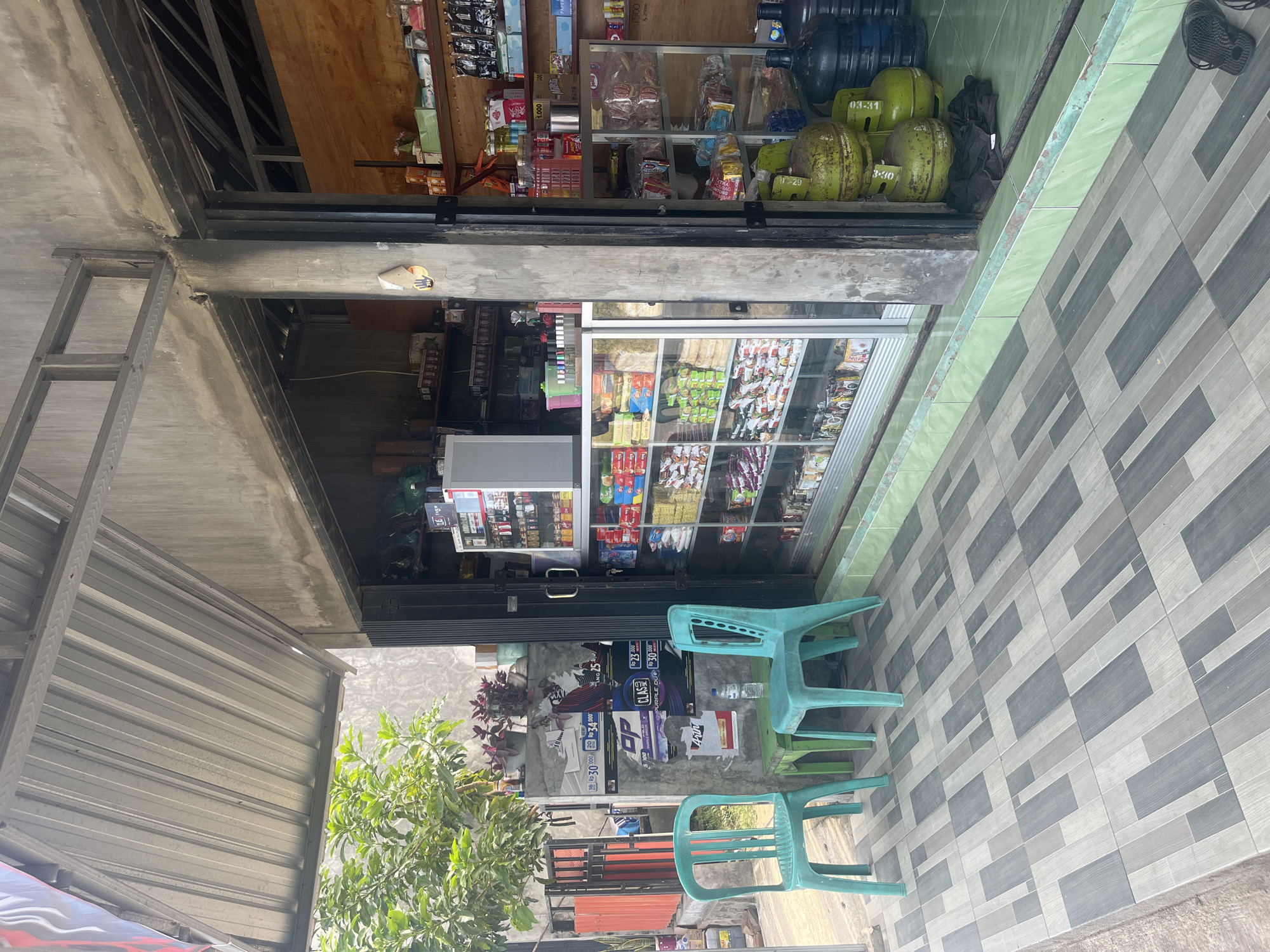 A storefront in Desa Lamunga, Indonesia is depicted in the image, featuring a small shop with an open-air display and a covered seating area. The shop is stocked with various goods, including bottled water, gas cylinders, and packaged food items, visible through glass display cases. Outside the shop, two turquoise plastic chairs and a green table are arranged on a tiled patio. Adjacent to the shop, a metal fence with orange accents and green foliage suggests a residential area or a garden. The overall scene portrays a typical local convenience store in a community setting.