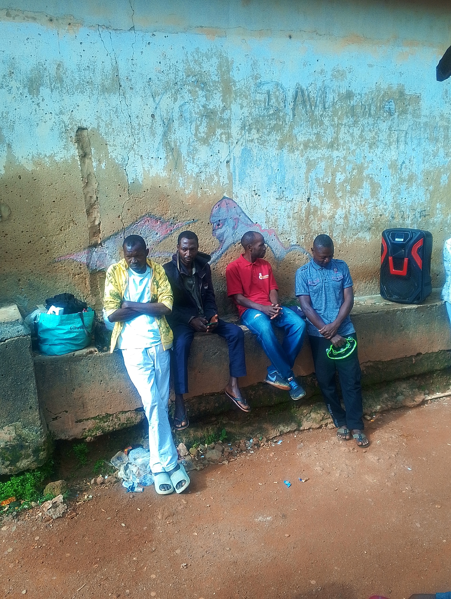 Four adult males are sitting on a concrete ledge in front of a textured, light blue and tan wall with graffiti. The man on the far left is wearing a yellow jacket over a white shirt and white pants, with his arms crossed. To his right, a man in a black jacket and dark pants is looking at his phone. Next to him, a man in a red polo shirt and blue jeans is looking down. The man on the far right is wearing a gray polo shirt and black pants, holding a green ring. A black and red portable speaker is to his right. A teal shopping bag is on the ground to the left of the first man, with some trash scattered on the ground in front of the ledge. The ground in front of the ledge is reddish-brown dirt.