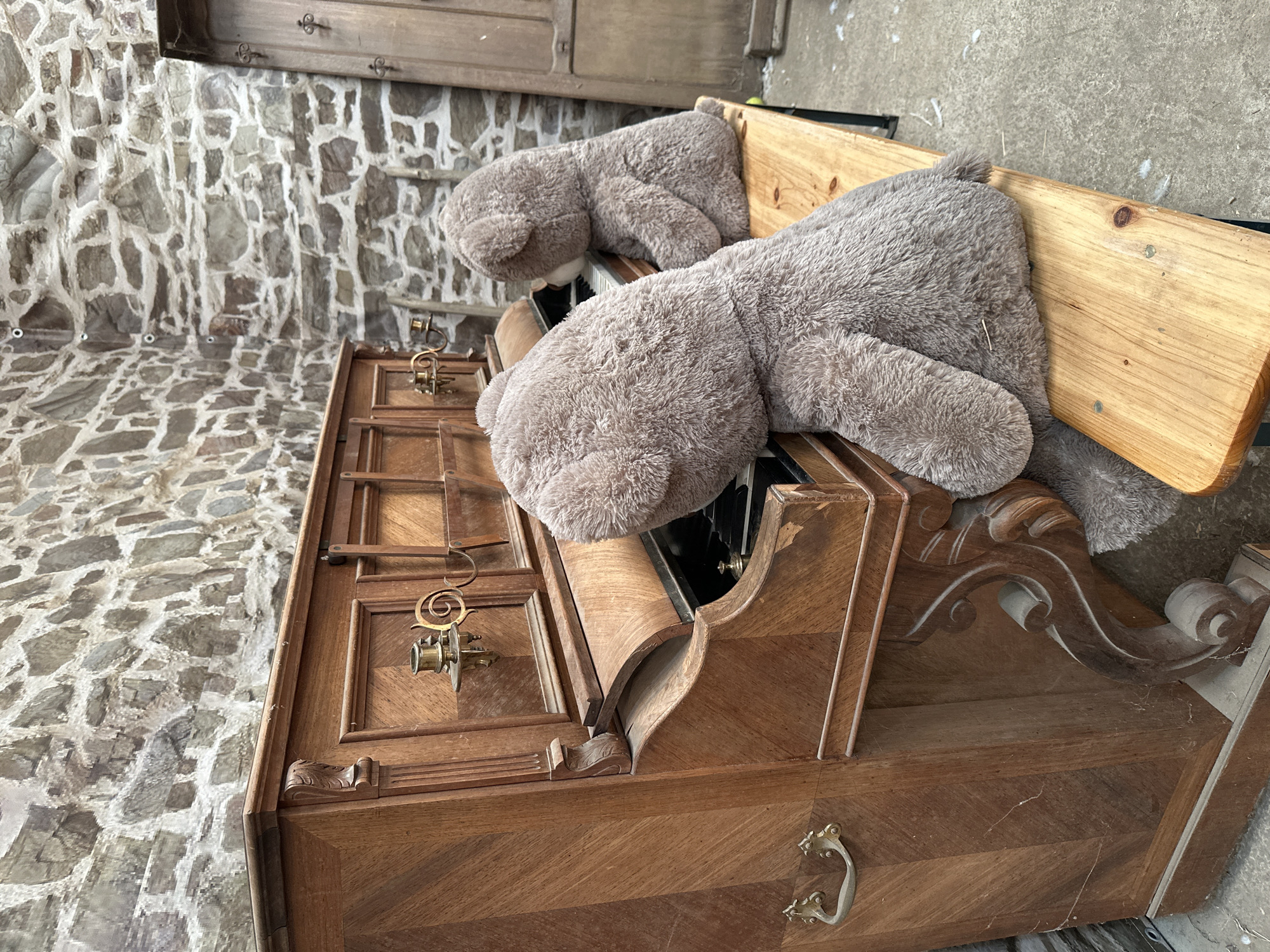 Two large teddy bears are seated on a wooden bench in front of an old, ornate wooden piano. The bear in the foreground is facing away from the camera, with its head angled towards the piano keys. The bear in the background is also facing the piano, with its head slightly turned. The piano is dark wood with intricate carvings and brass candle holders. The scene is set against a rustic stone wall, with a wooden door visible in the background. This image was taken in Gonneville-la-Mallet, France.