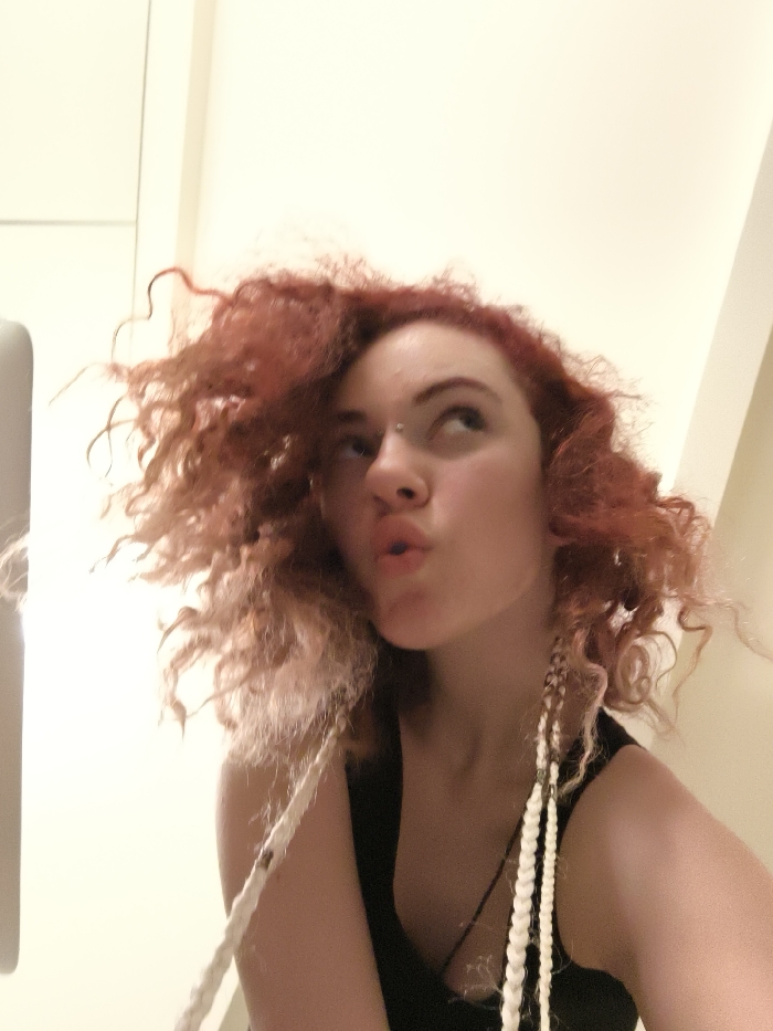 The image displays a young female, framed from the chest up, in an indoor setting. Her hair is voluminous, reddish-brown, and curly, with several light-colored, braided or dreadlock-style extensions hanging on both sides of her head. A small, metallic barbell piercing is visible between her eyebrows. Her lips are pursed in a pouting expression, and her eyes are directed upwards and slightly to the right of the frame. She is wearing a dark, sleeveless top, with a dark pendant necklace faintly visible on her chest. The background consists of plain, light-colored walls, indicative of a generic interior space. There are no discernible architectural features, landmarks, or other visual cues within the image to identify a specific location within Paris, France, or any other city.