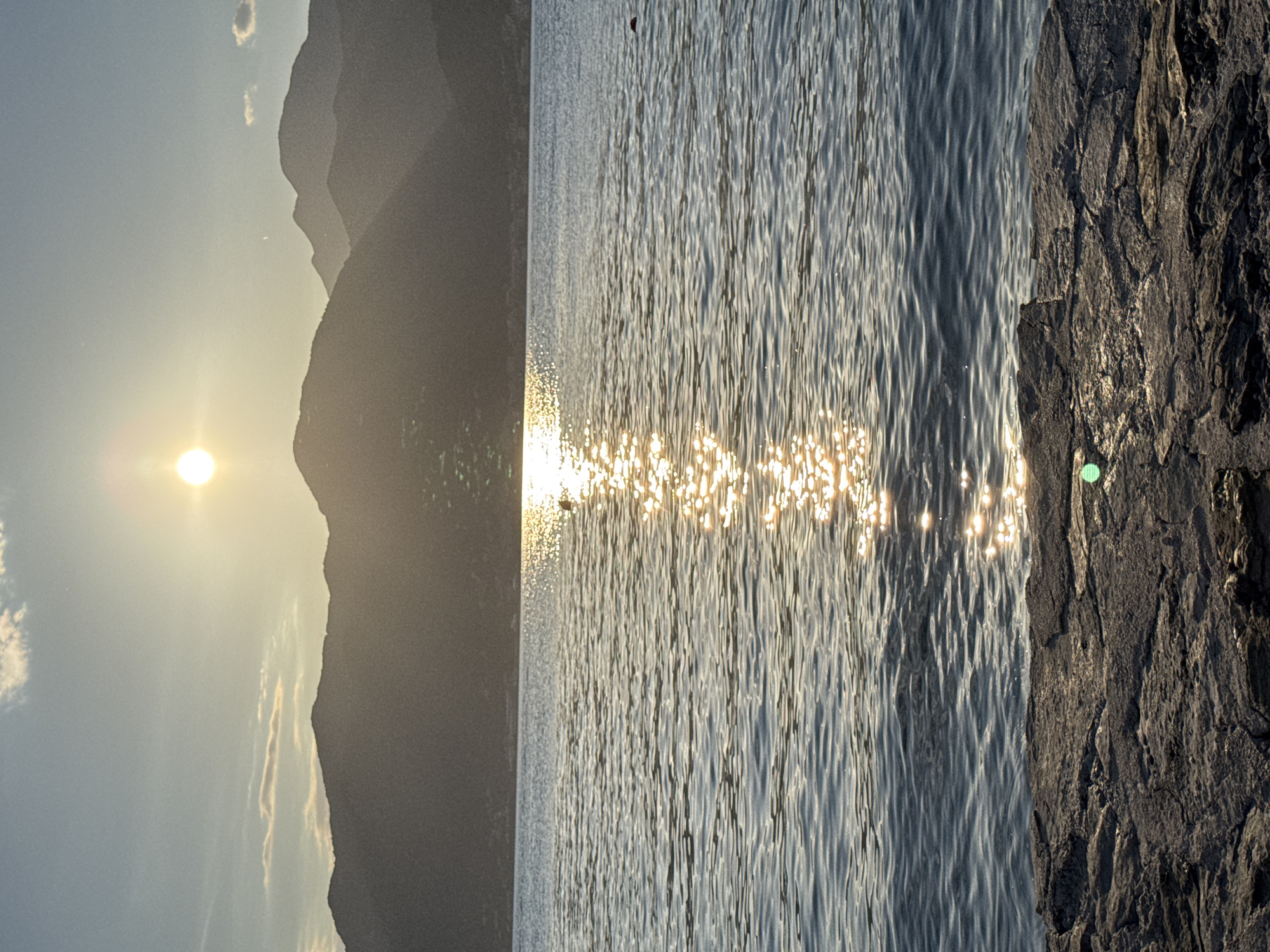 This media file depicts a serene natural landscape scene at sunset. The image is oriented vertically, with the foreground on the right side occupied by a rough, textured rock formation. Adjacent to the rocks, a body of water, likely a sea or lake, fills the middle portion of the frame. The water's surface is rippled, reflecting the warm, golden light of the setting sun, creating a dazzling, shimmering effect across the water.
In the background, on the left side of the image, silhouetted mountain ranges recede into the distance, their dark forms contrasting with the bright sky. The sun, a distinct orb of light, is visible in the upper left quadrant, casting a soft glow and contributing to the atmospheric lighting of the scene. Wispy clouds are scattered across the sky, some tinged with the sunset colors.
The overall mood is peaceful and tranquil, evoked by the natural beauty of the sunset over water and mountains. There are no people or visible objects that suggest a specific activity, aside from the implicit presence of the natural environment. The time of day is clearly sunset, indicated by the low angle of the sun and the warm, golden light. The location cues suggest a coastal or lakeside setting with mountainous terrain, consistent with Donja Lastva, Montenegro. There is no visible text in the image.