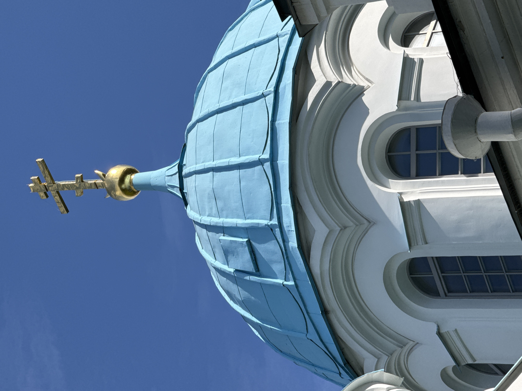 A close-up, low-angle shot captures the turquoise dome and golden cross of an Orthodox church against a clear blue sky in Saint Petersburg, Russia. The dome is segmented and weathered, showing lines of construction. A multi-tiered golden cross, a symbol of Orthodox Christianity, crowns the dome, perched atop a golden sphere. The white building below features arched windows with dark panes, set within decorative white architectural elements. The image is a detail of the upper portion of a religious structure, emphasizing its architectural and symbolic features.