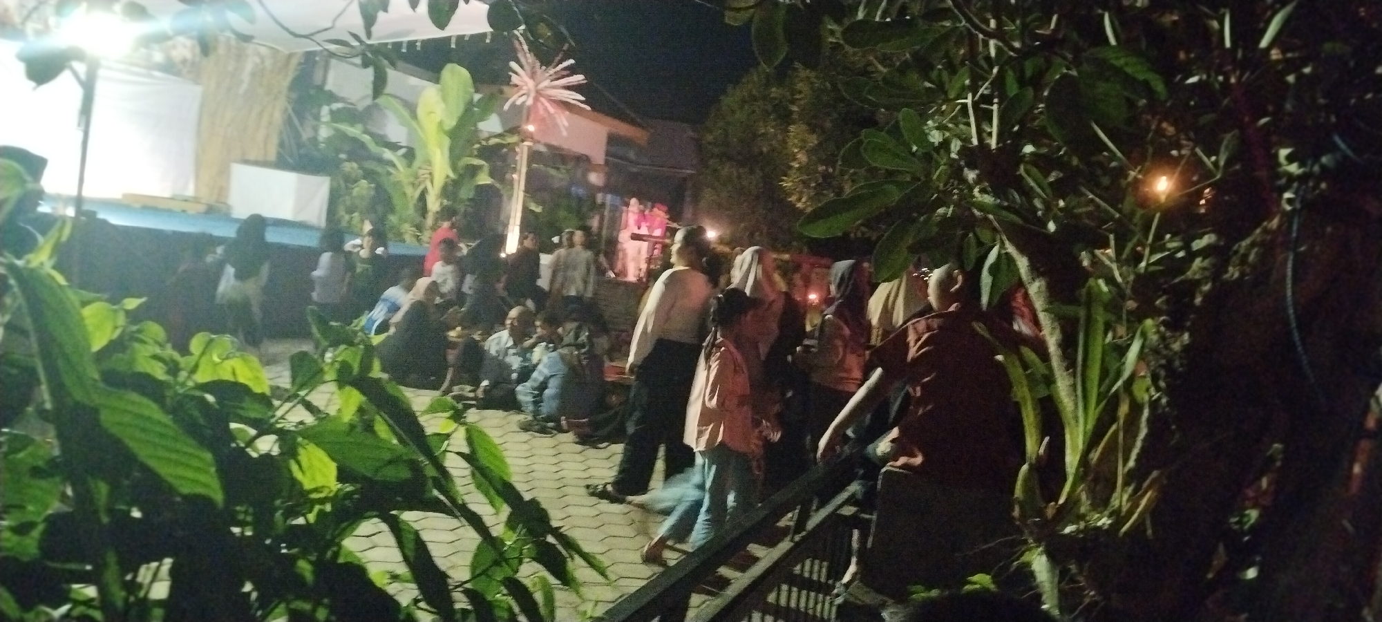 The image depicts an outdoor evening gathering in Keting, Indonesia. A group of people are seated and standing on a paved area, possibly a courtyard or public space, with lush green foliage in the foreground and framing the scene. In the background, a white screen is visible, suggesting a projection or performance area. A decorative, star-like light fixture hangs above the gathering, and ambient lighting illuminates the scene. The people are casually dressed and appear to be engaged in conversation or observing an event. The overall atmosphere is relaxed and communal.