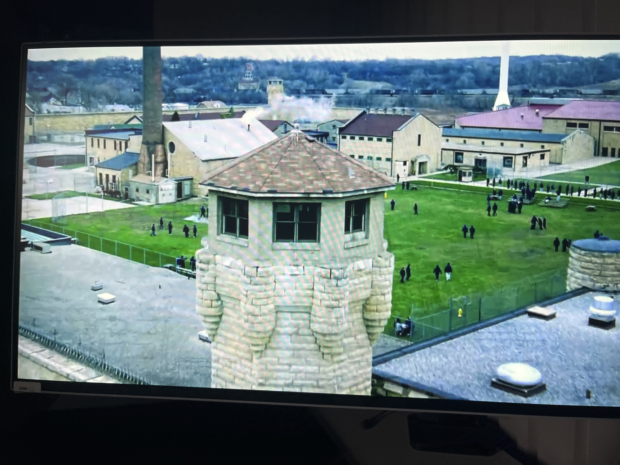 The image shows an aerial view of a prison complex. A central watchtower, constructed from light-colored brick with turrets and a tiled roof, is in the foreground. Beyond the watchtower, various prison buildings with tan walls and different colored roofs are visible. A large, grassy courtyard is occupied by numerous small, dark figures, presumably inmates, some walking individually or in small groups, while others appear to be gathered in larger formations. A fence line, possibly an outer perimeter, encloses parts of the complex. The background reveals a more distant landscape with trees and industrial-looking structures, including a tall smokestack. The overall scene suggests a correctional facility with an active population. There is no indication of the city of Varna, Bulgaria in this image.
