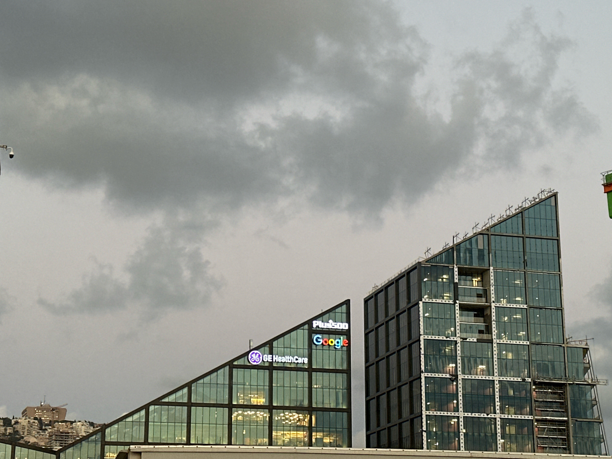 The image shows a cloudy sky above a modern building complex in Haifa, Israel. On the left, a triangular building features prominent illuminated signs for "Plus500" and "Google" at its apex, and a "GE HealthCare" logo further down the facade. The building has a glass facade with a grid-like structure. To the right, a taller, rectangular building also exhibits a glass facade with a similar grid pattern, appearing to be under construction or renovation with scaffolding visible on its side. In the background, on a hill, there are several multi-story buildings with lights on, indicating an urban setting. A security camera is visible on a pole in the upper left corner of the image.