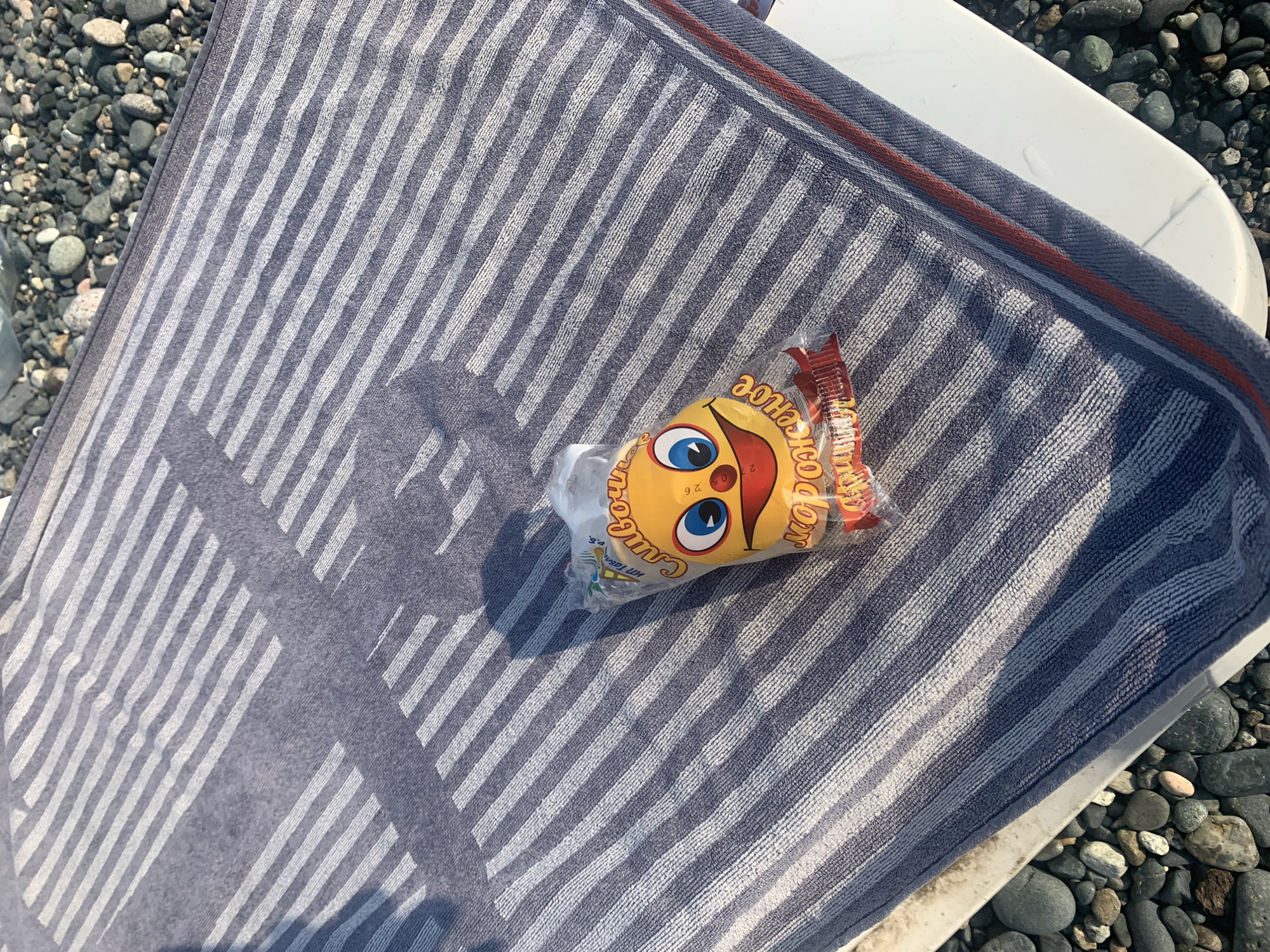 An overhead, high-angle shot captures a beach towel with alternating navy blue and white stripes, laid on a white surface next to a collection of smooth, dark grey pebbles. A transparent plastic bag containing a yellow, smiley-face-shaped candy wrapper rests on the towel. The wrapper is partially visible, revealing the design of a face with large blue eyes, a red mouth with a tongue sticking out, and text in Cyrillic script that appears to be "Сливочное" (Slivochnoye) and "Мороженое" (Morozhenoye), suggesting the candy is ice cream flavored. The shadow of an unseen object falls across the towel, indicating bright sunlight. The scene is set outdoors, likely at a beach in Gulripshi, Abkhazia.