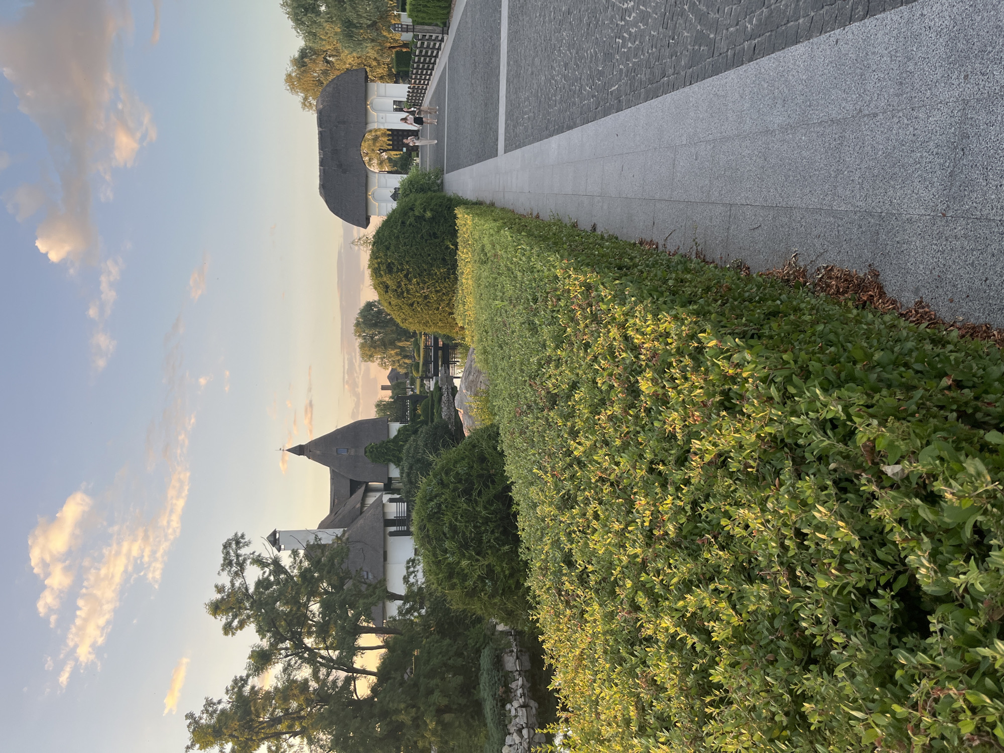 The image depicts a scenic pathway with manicured hedges and cobblestone paving, leading towards a thatched-roof archway. In the background, several buildings with similar thatched roofs are visible, nestled amongst trees and greenery. The setting appears to be a park or resort area, possibly within the city of Dnipro, Ukraine, given its architectural style and landscaping. The sky is partly cloudy with the soft hues of sunset casting a warm glow on the scene. Three individuals are seen walking on the pathway near the archway, engaged in a leisurely stroll. The overall atmosphere is serene and picturesque, suggesting a peaceful outdoor environment.